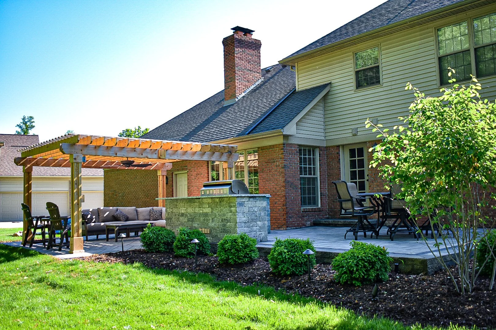 Backyard with outdoor seating area, pergola, brick fireplace, patio, lush greenery, and the back of house with multiple windows and gable roof.