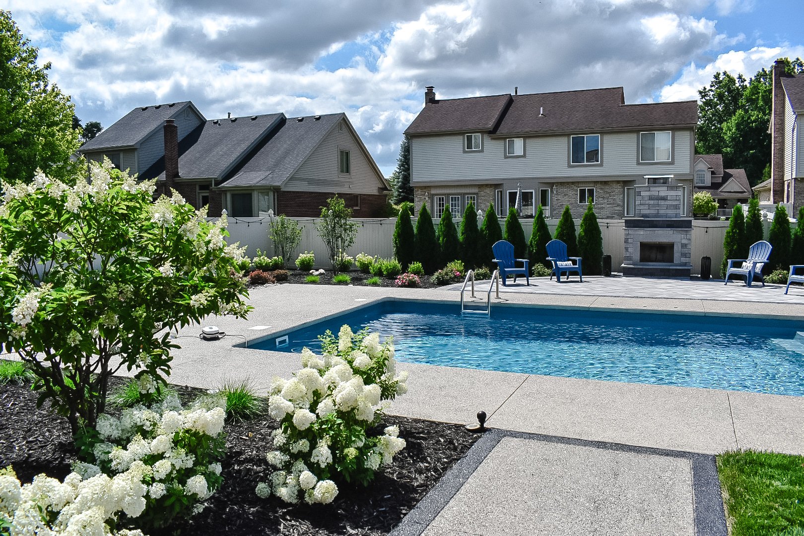 Residential backyard with swimming pool, surrounding plants and flowers, blue Adirondack chairs, stone fireplace, and neighboring houses under a partly cloudy sky — GLC Lawn & Landscape