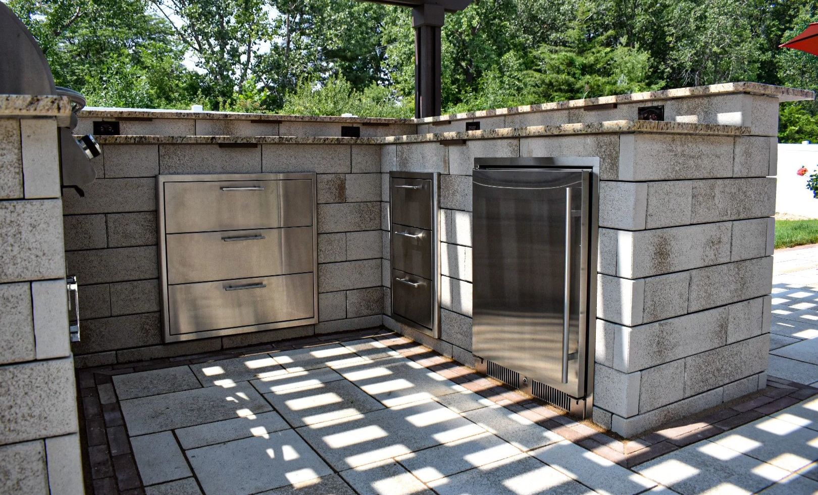 Outdoor kitchen with stone brick counter, stainless steel drawers, storage cabinets, and a mini refrigerator, surrounded by greenery under open sky.
