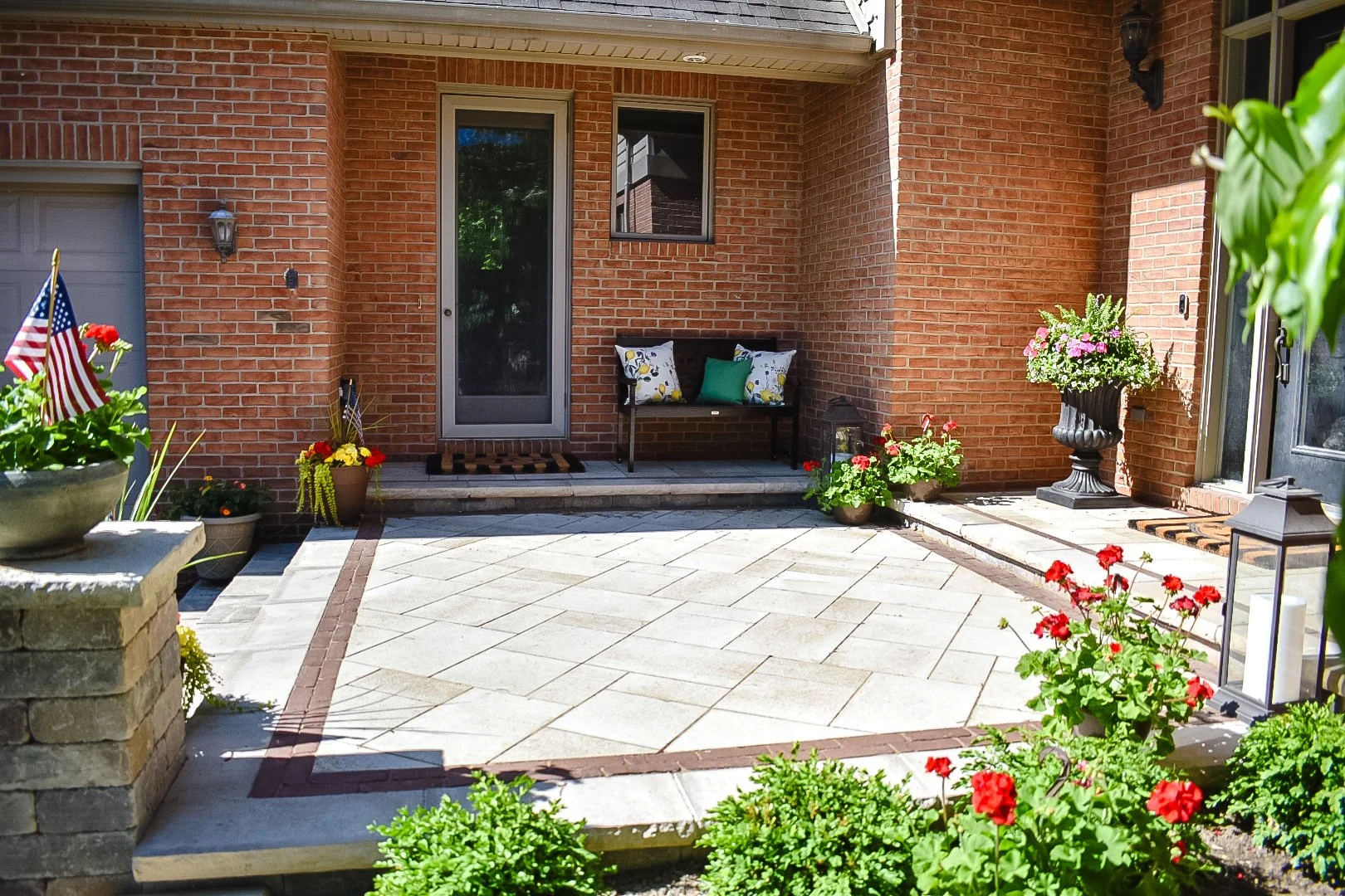 A brick house porch with decorative potted plants, flowers, and a black bench with cushions, with a glass door and a window.