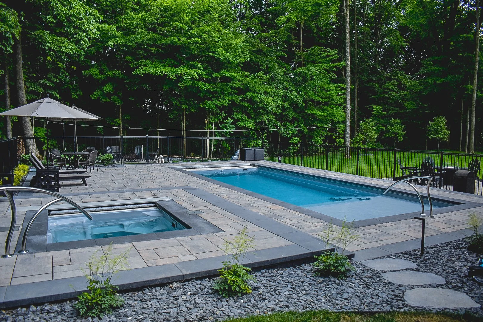 Backyard with a swimming pool and hot tub, surrounded by a patio with outdoor furniture and lush green trees in the background.