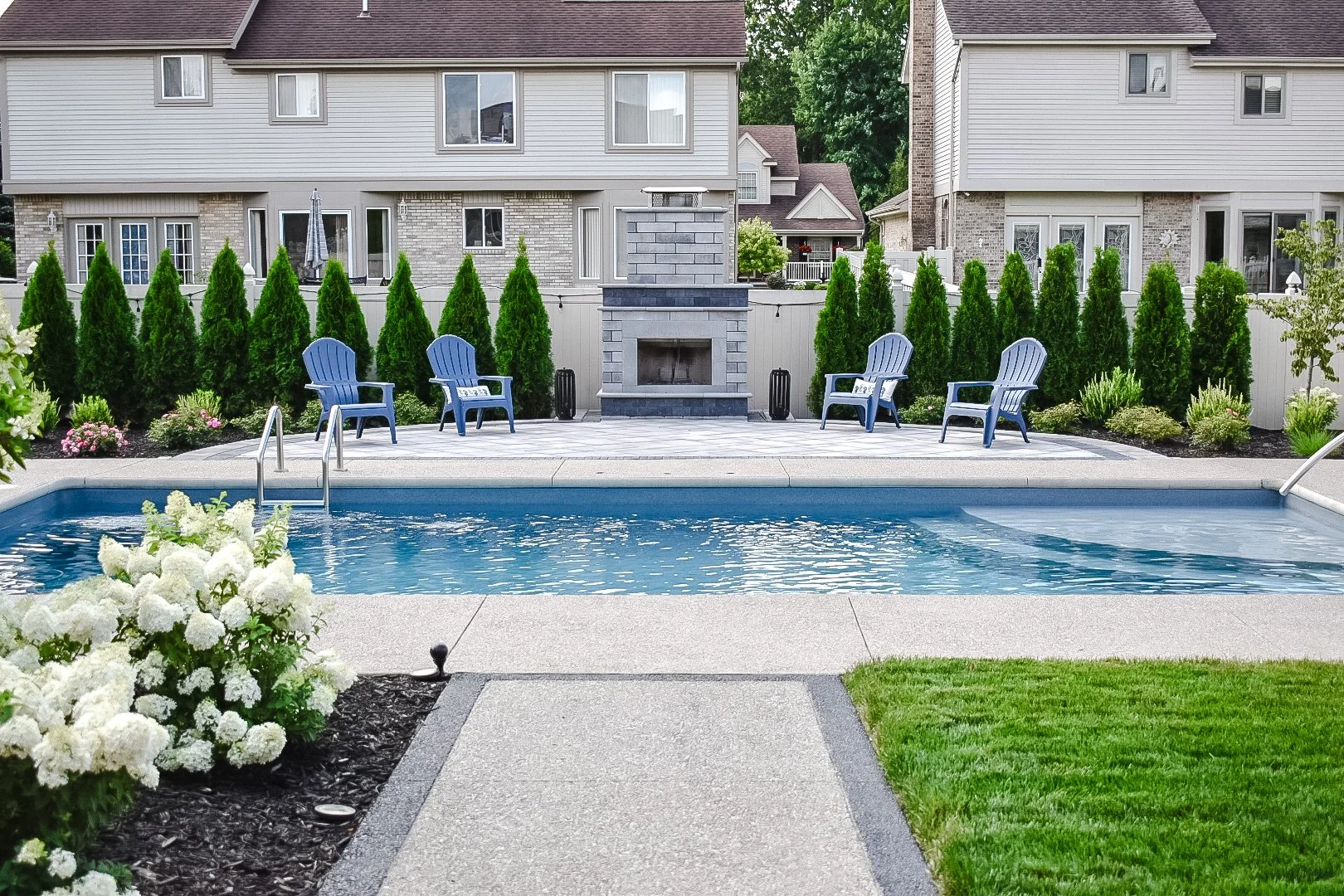 A backyard with a swimming pool, surrounded by a concrete deck and green grass, with four blue chairs, a stone fireplace, and lush green bushes against a fence, with residential buildings in the background.