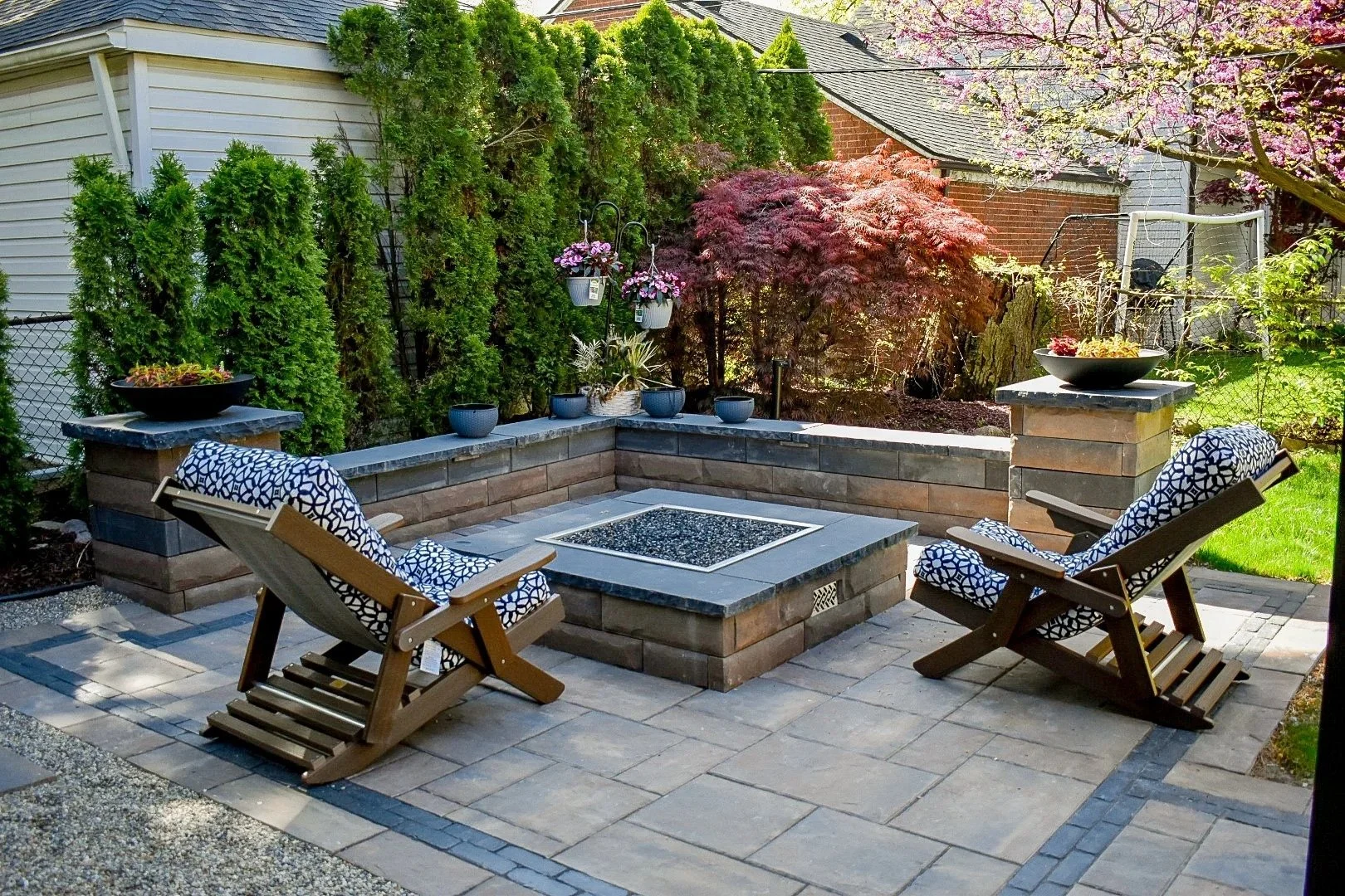 A backyard patio with two wooden lounge chairs with blue and white patterned cushions, a built-in stone fire pit, and various potted plants. Green trees and a house with a gray roof are in the background.