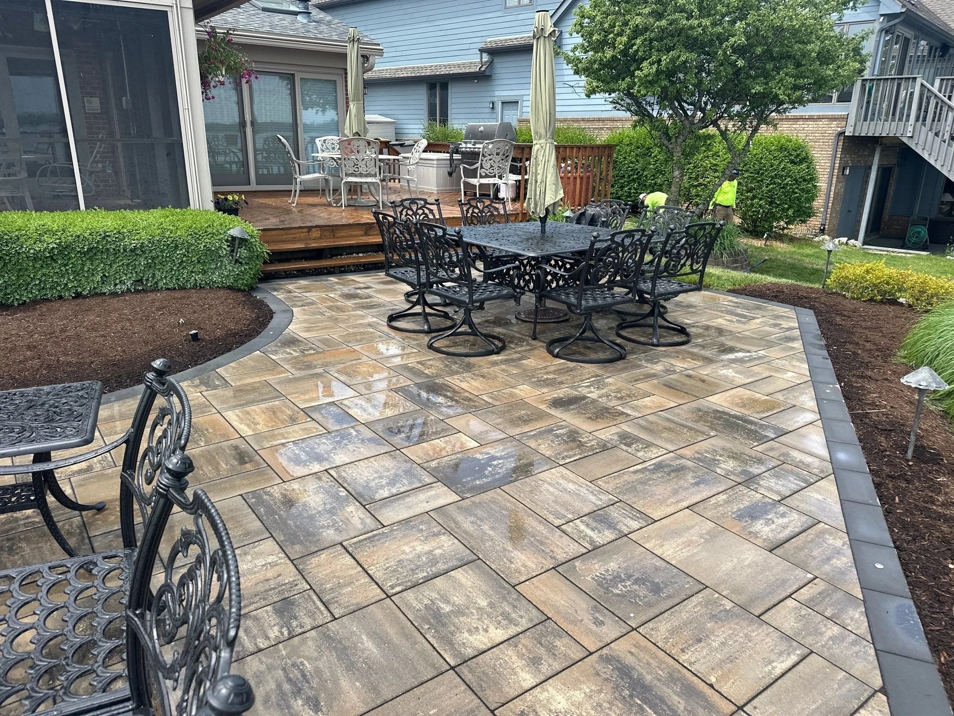 Backyard patio with black wrought iron dining table and chairs, wooden deck with white outdoor furniture and grill, lush green bushes, trees, and a blue house in the background, during rainy weather.
