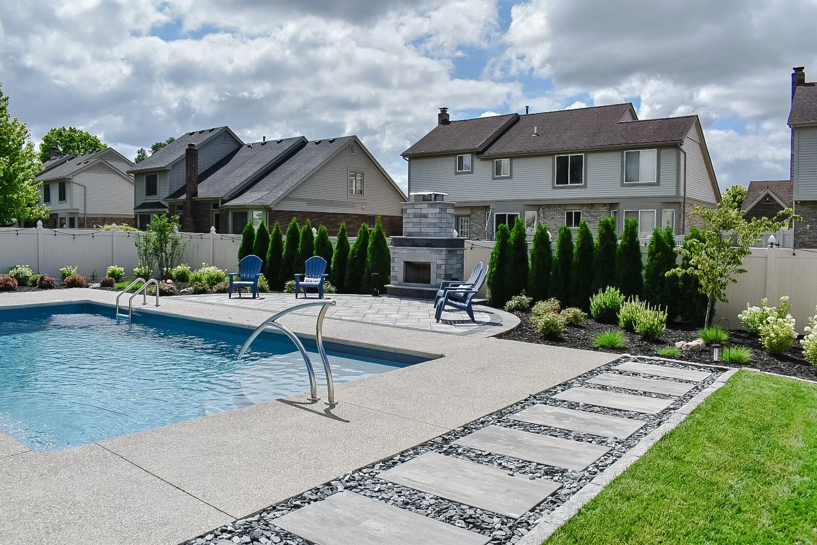 Backyard with swimming pool, patio seating, landscaped garden, and neighboring houses under partly cloudy sky.