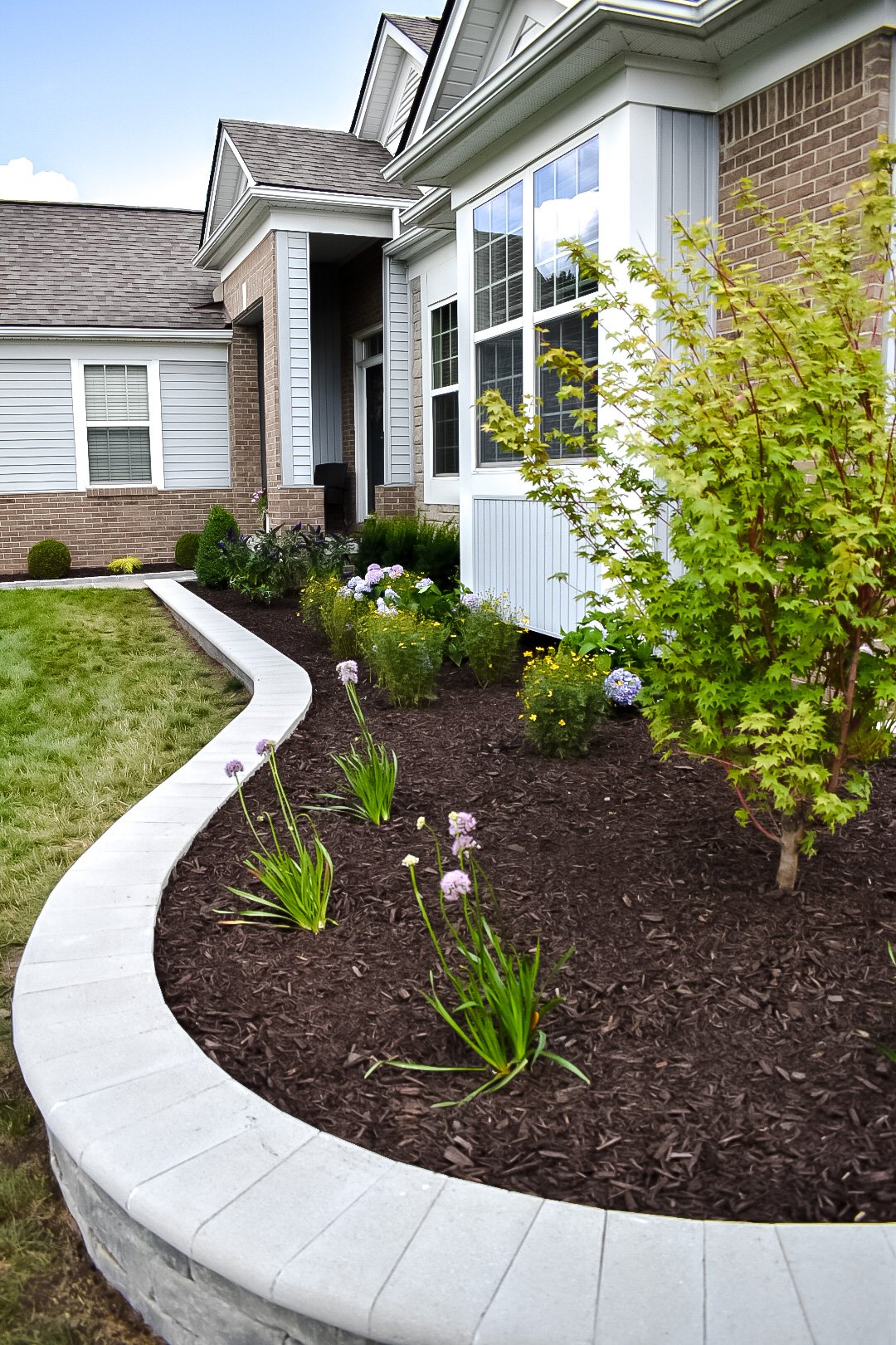 Front yard with flower bed and mulched plants next to house, featuring brick and siding exterior with large windows, a lawn, and a curved white border around the flower bed.