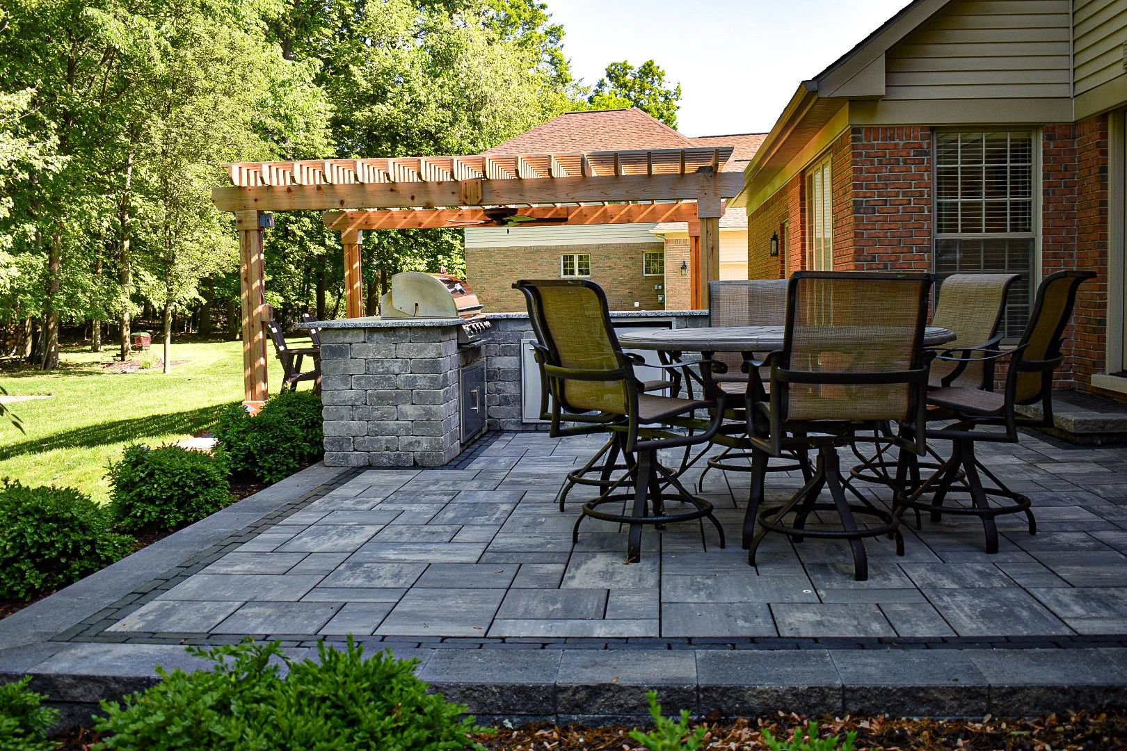 A backyard patio with a brick house, a stone outdoor kitchen with a built-in grill, a wooden pergola, and a large round table with six chairs. There is greenery and trees in the background.