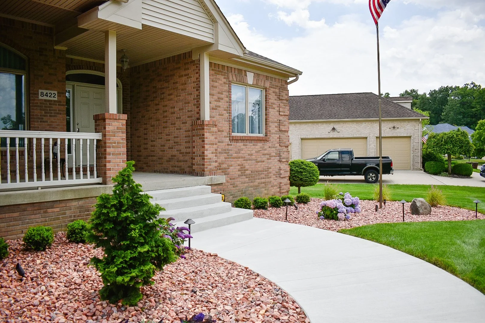 Front yard and entrance of a brick house with well-kept gardens and a concrete walkway leading to the front stairs. In the background, a black pickup truck is parked in front of a detached garage with beige doors.