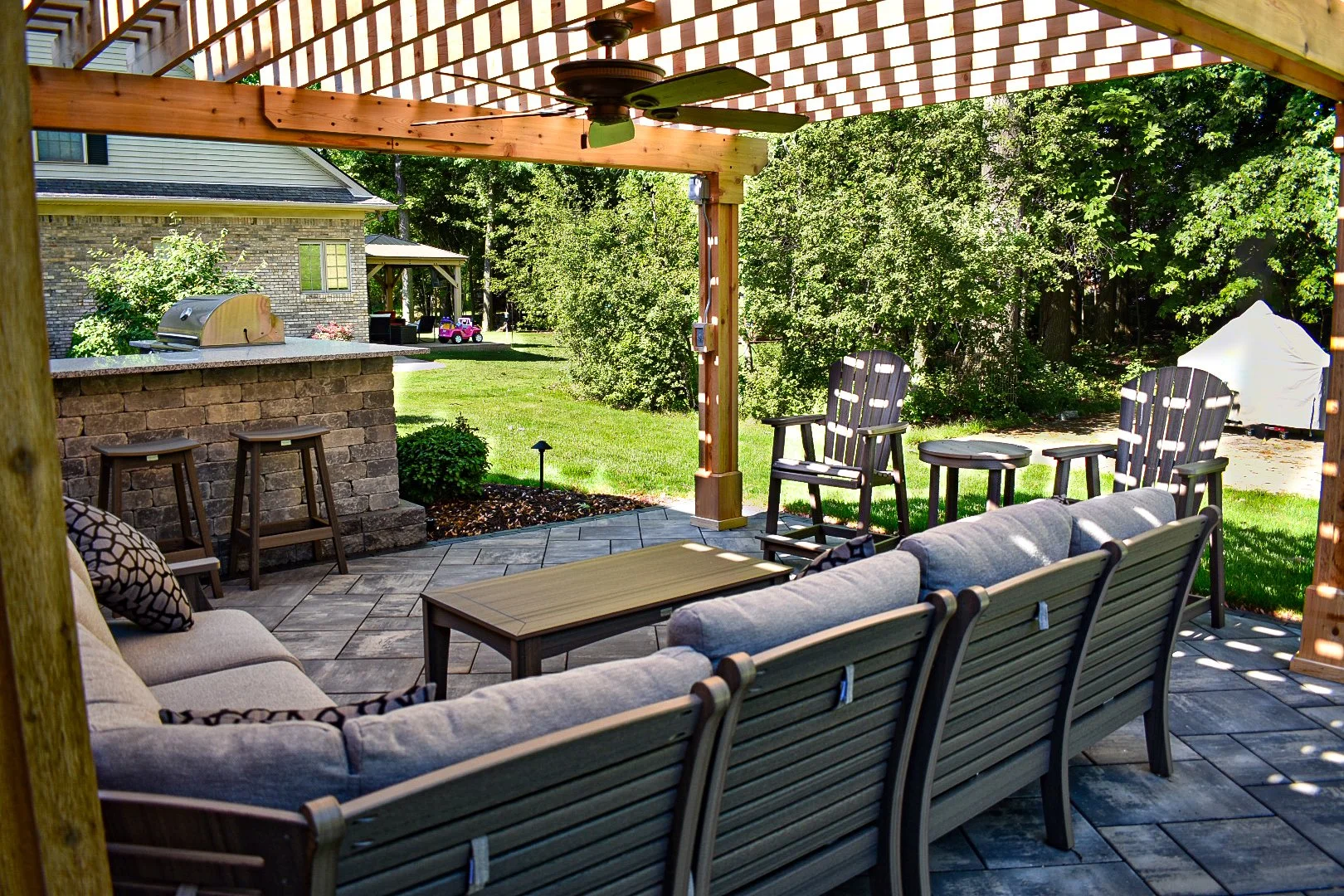 Outdoor patio with seating area, including a cushioned sofa, Adirondack chairs, and a coffee table. There is a stone outdoor kitchen with a built-in grill, a bar counter with stools, and a ceiling fan. The patio is covered by a pergola with a lattice