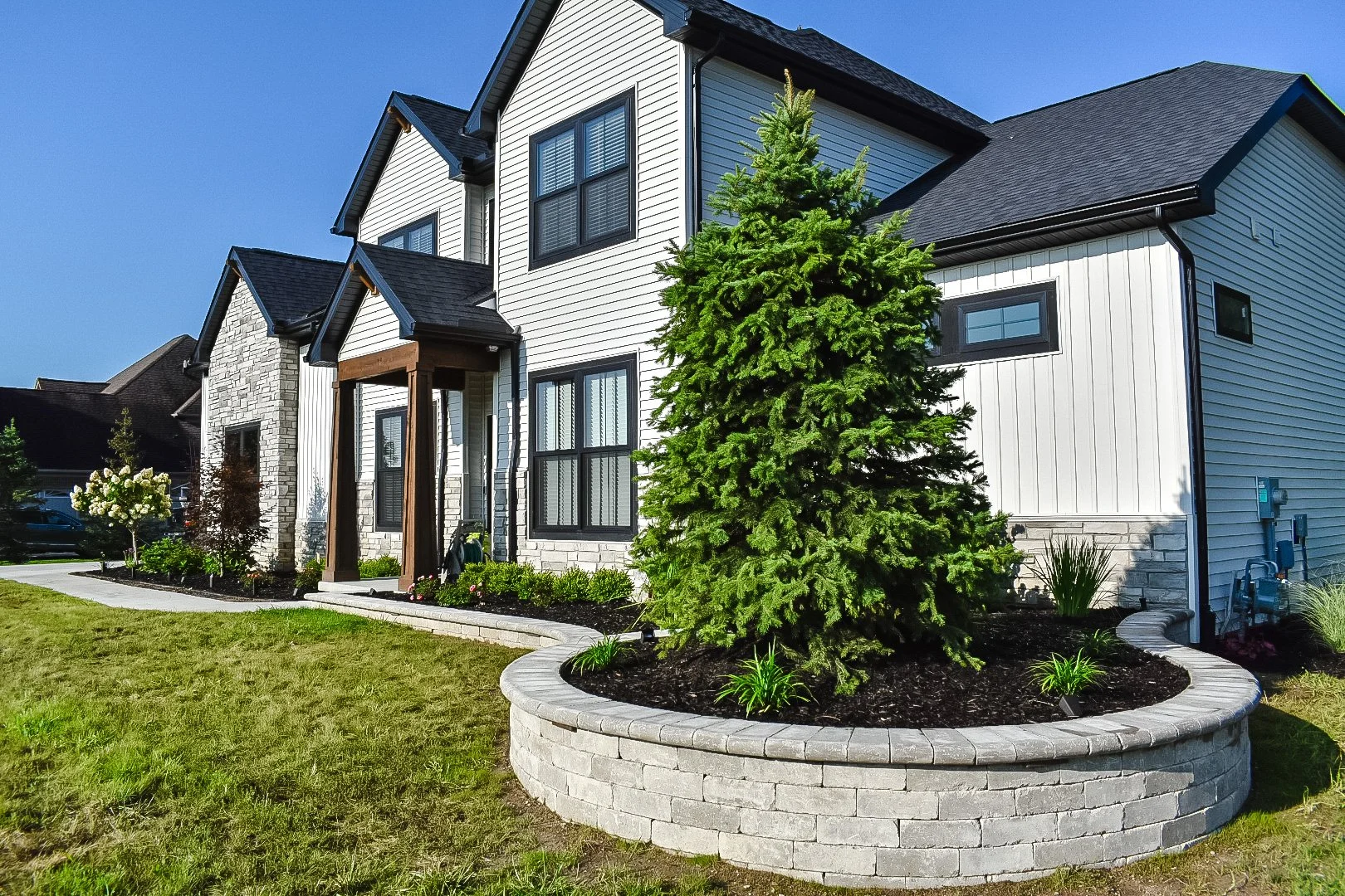 Modern two-story house with white siding and black trim, landscaped front yard, and a large evergreen tree in a circular brick planter.