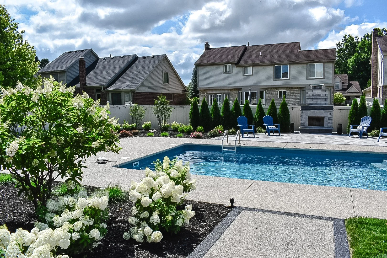 Backyard with a swimming pool, patio chairs, a fireplace, landscaping with shrubs and trees, and neighboring houses in the background under a partly cloudy sky.