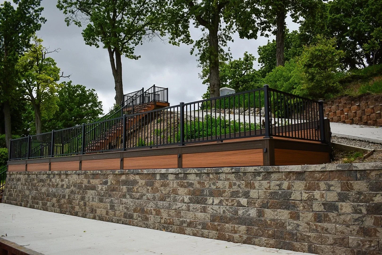 A multi-level landscape terrace with a stone retaining wall, wooden siding, black metal railing, and a concrete pathway, surrounded by green trees and shrubs.