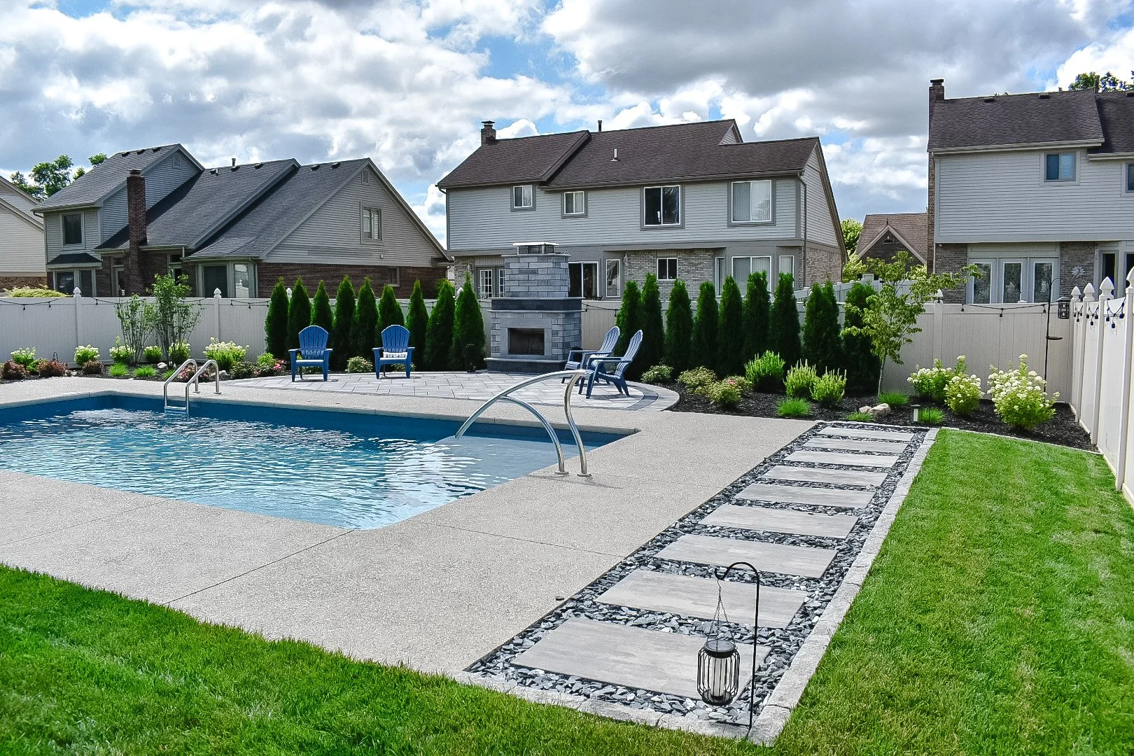 Backyard with swimming pool, blue chairs, fire pit, garden, pathway, and neighboring houses.