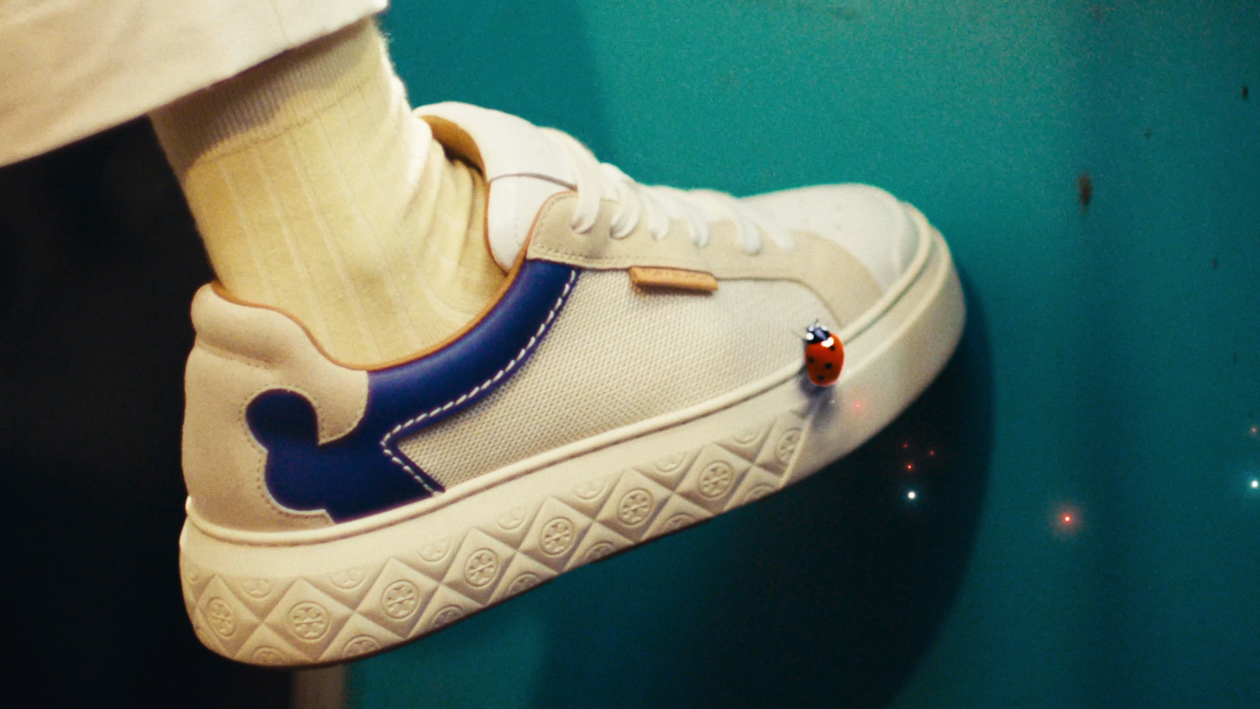 Close-up of a person’s foot wearing a white sneaker with navy and beige detailing, beige socks, and beige pants. A small red ladybug figurine is attached to the sneaker’s side, connected to a tiny red light that glows. The background is a teal-colore