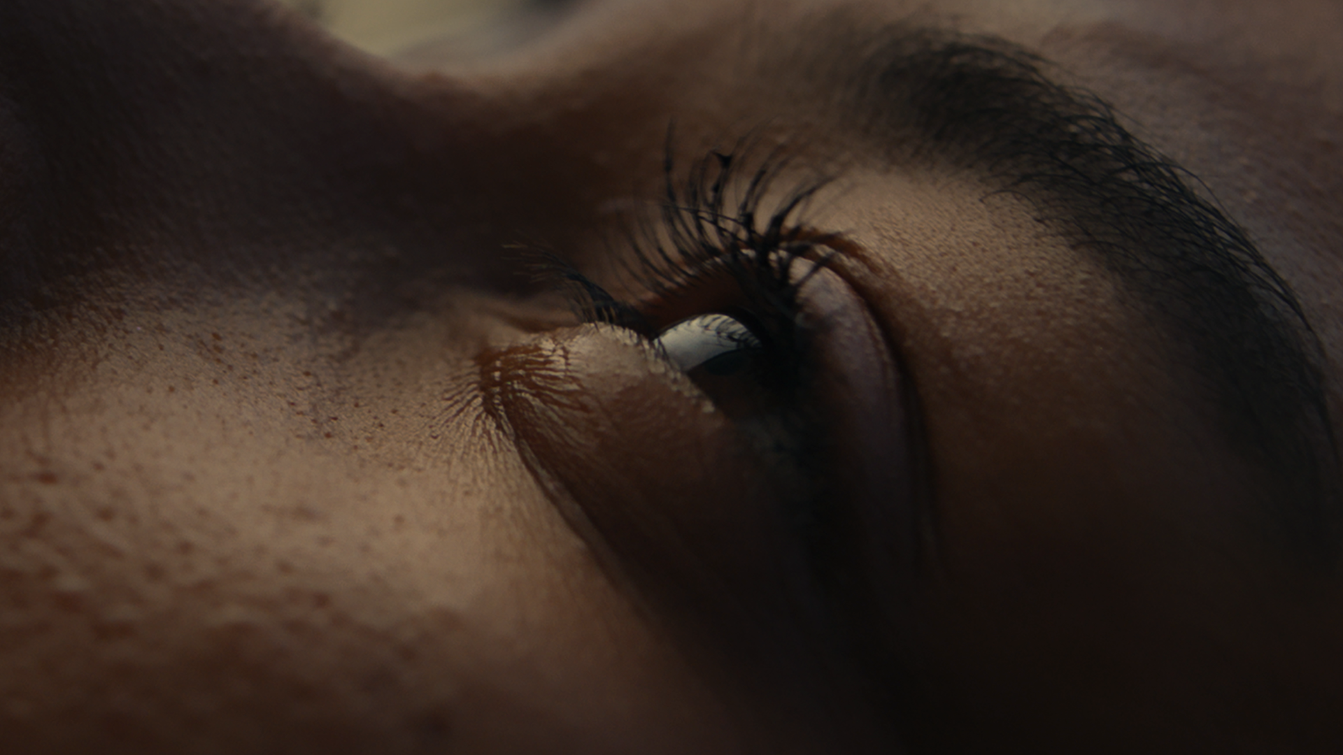 Close-up of a person's eye, showing detailed eyelashes, eyelid, and surrounding skin in soft lighting.