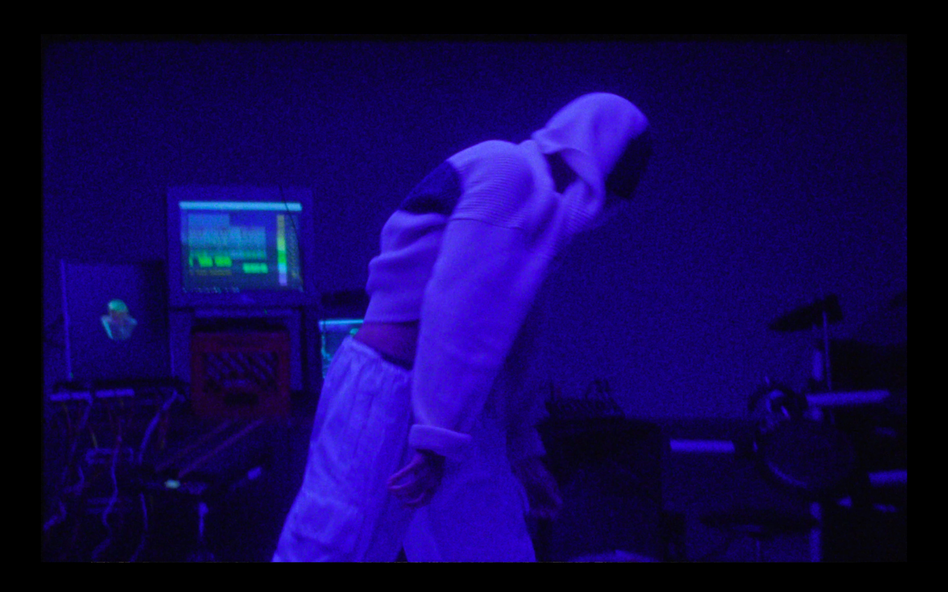 A person in a hoodie and baggy pants bowing in a dimly lit room with neon blue lighting, computers, and a bicycle.