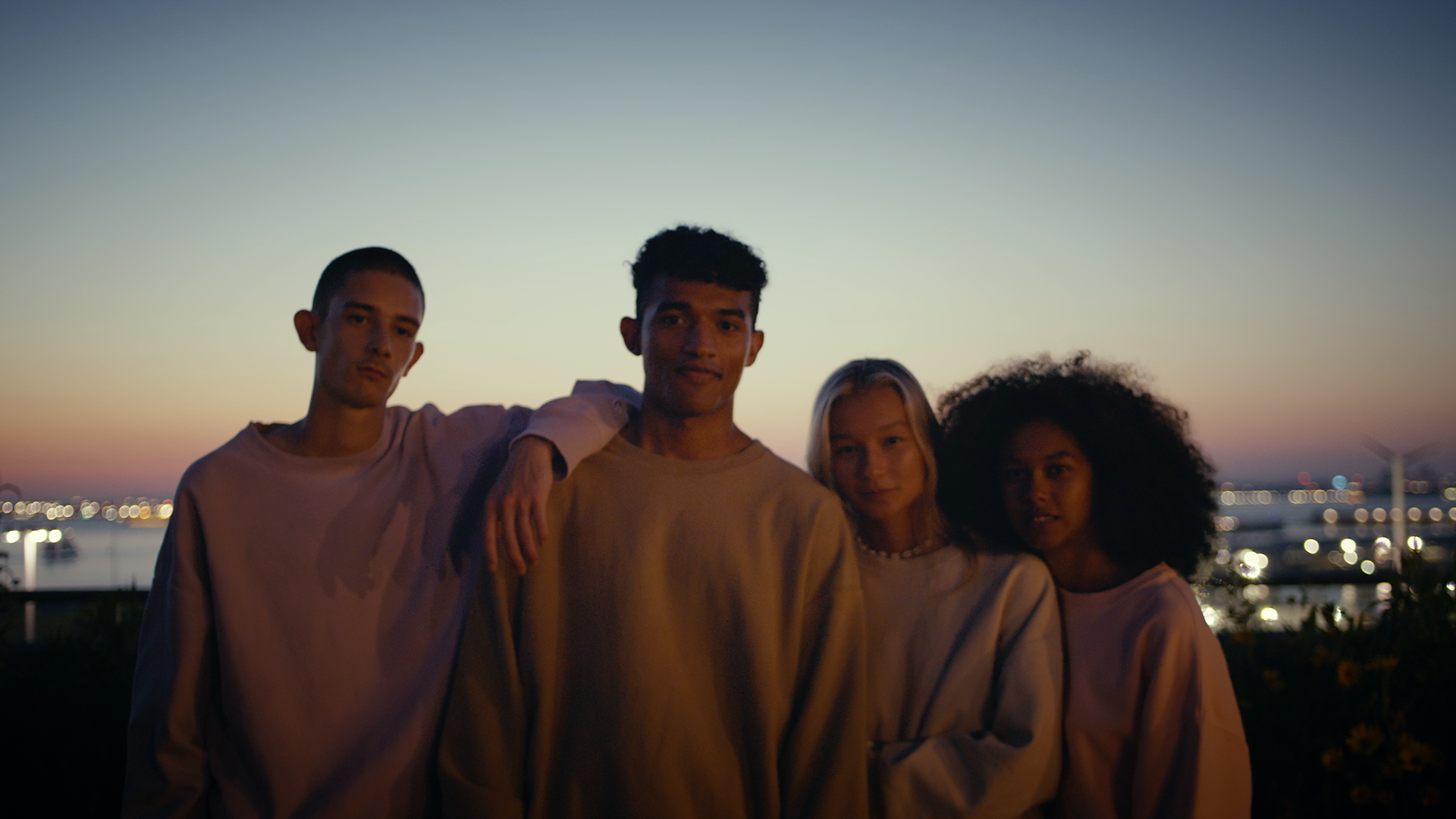 Four young people standing together outdoors at sunset with city lights and water in the background.
