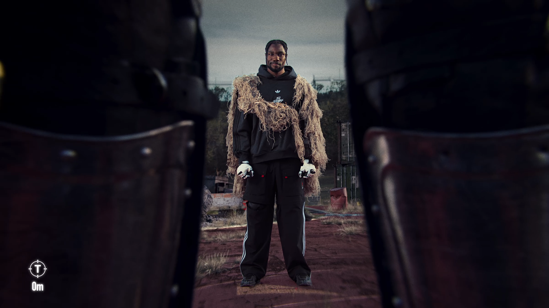 A man standing outdoors on a dirt surface, viewed through the gap between a pair of dark shoes in the foreground, with a cloudy sky and trees in the background.
