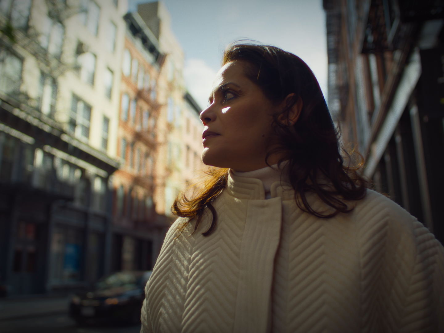 Profile of a woman with brown hair in a cream-colored jacket, standing outdoors in a city street with buildings in the background, in bright sunlight.