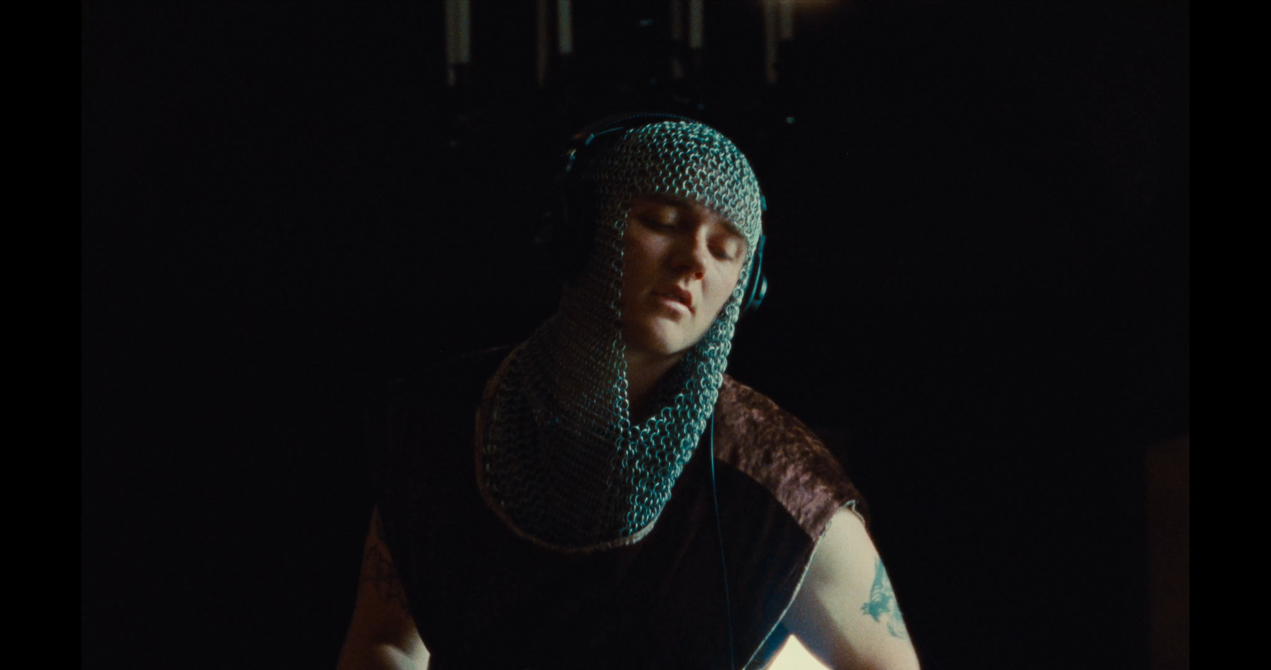 A person wearing chainmail protective headgear and headphones, with closed eyes, in a dark environment.