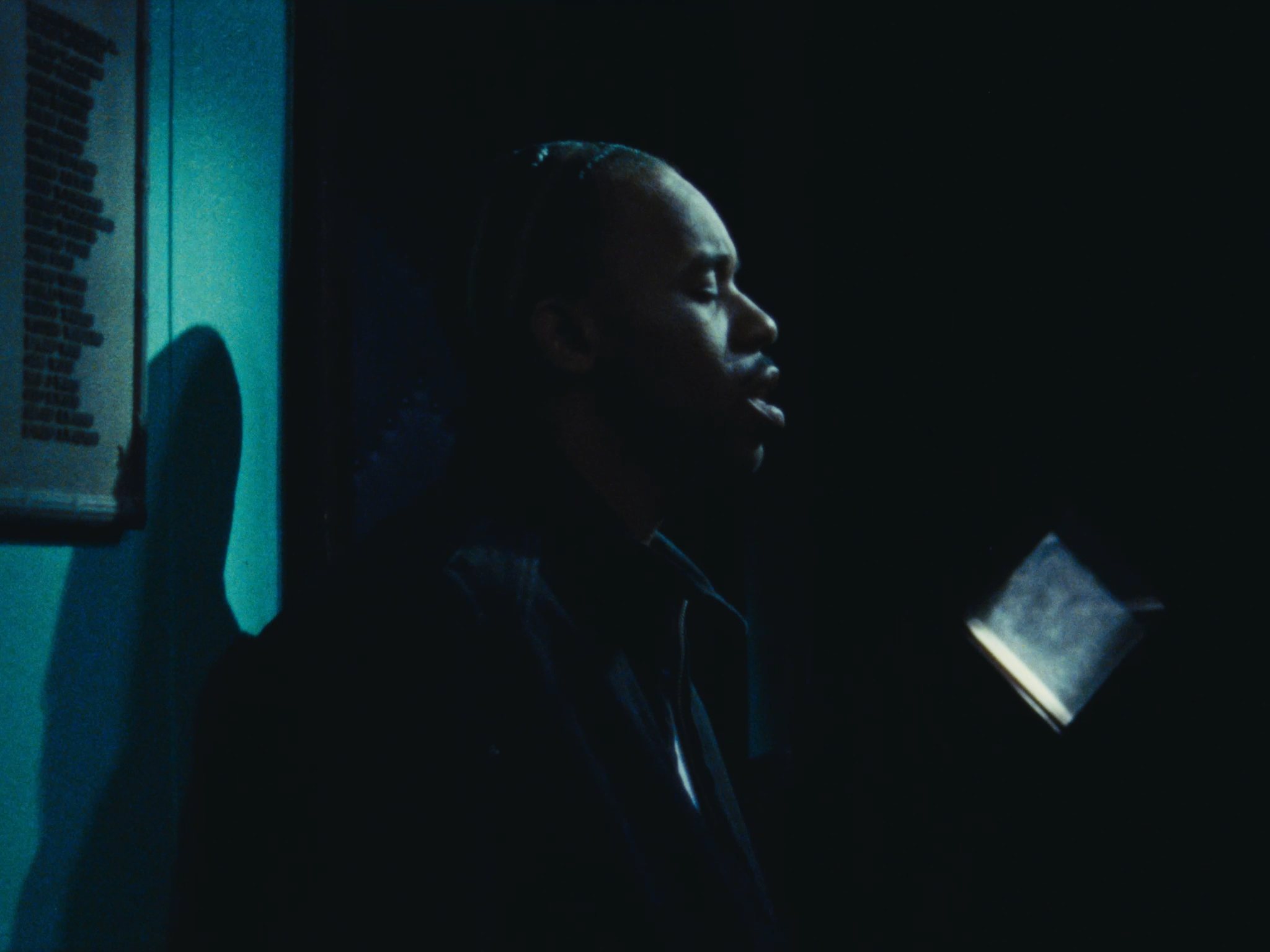 A person with braided hair and closed eyes, standing in a dark room with faint light illuminating their face.