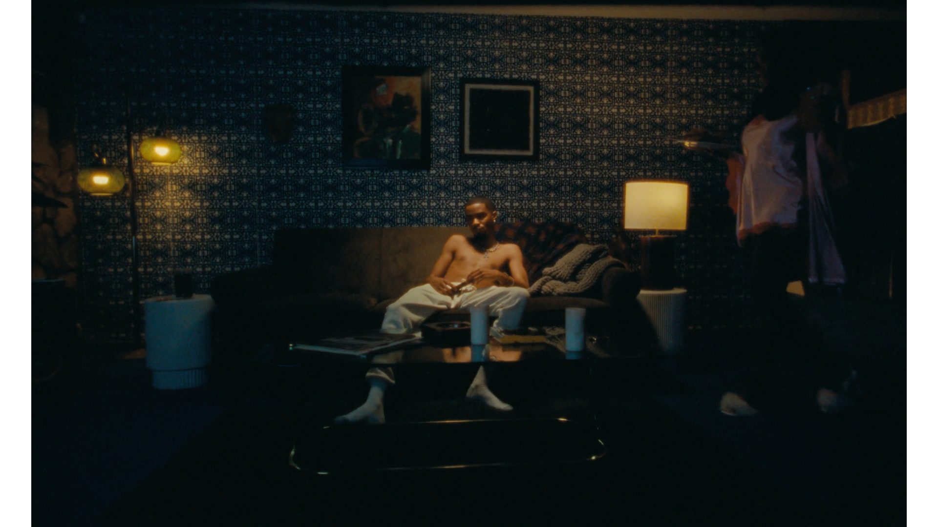 A young man with dark skin sits shirtless on a brown sofa in a dimly lit room. He wears white pants, a pearl necklace, and has a cross necklace around his neck. In front of him is a glass table with candles and magazines. The room has dark patterned 
