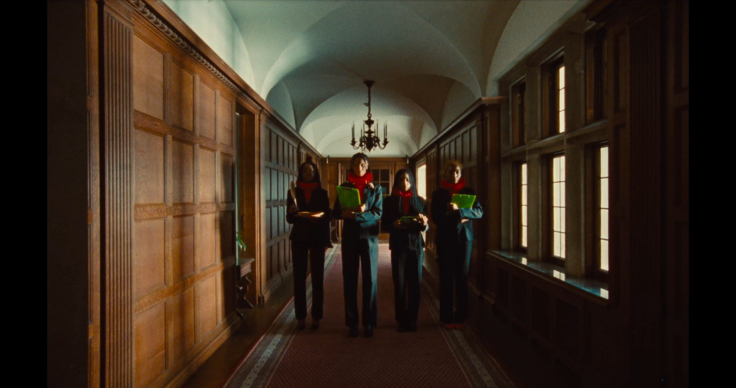 Four women dressed in black suits and red scarves walking down a corridor with wood-paneled walls and large windows, carrying attaché cases and folders, with a chandelier hanging from the arched ceiling.