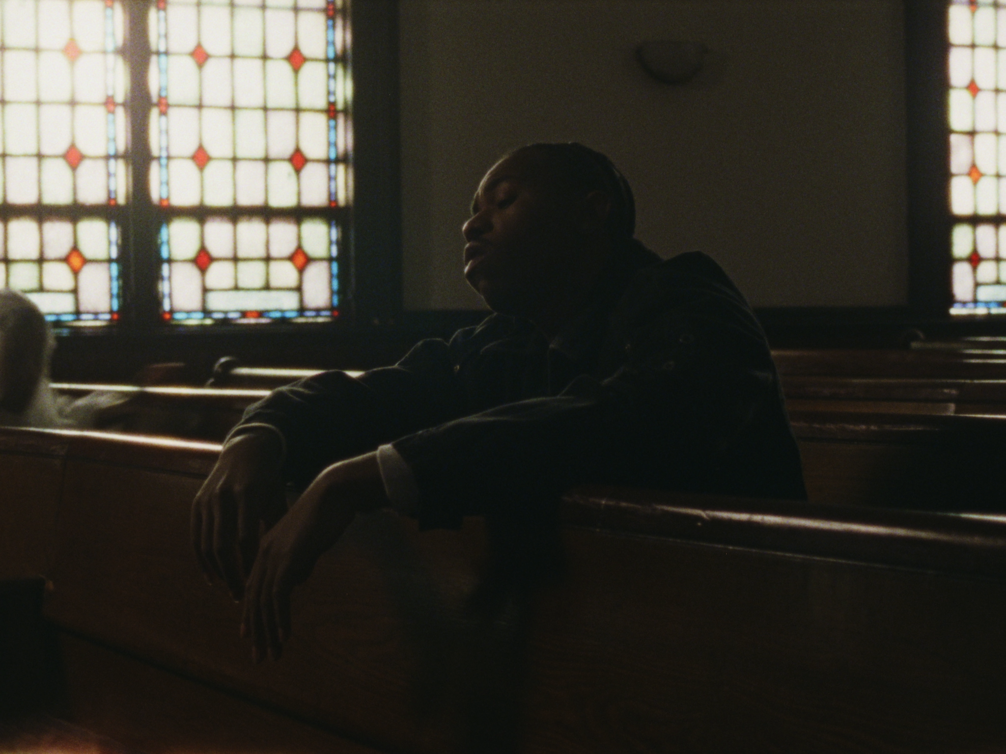 A person sitting on a church pew in front of stained glass windows, with their eyes closed and head tilted back in a reflective pose.