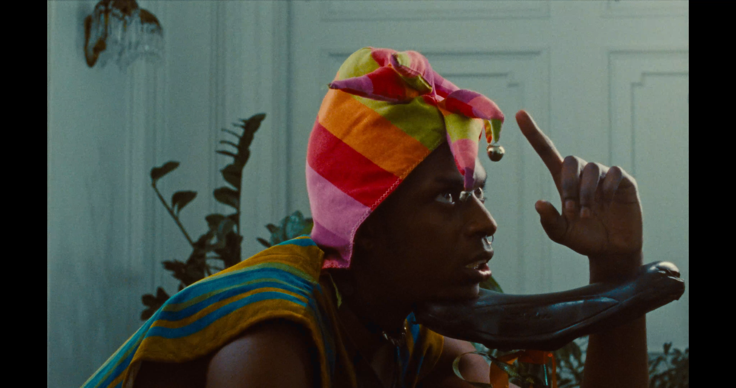 A person wearing a colorful striped headwrap and a striped yellow and blue top gestures with their right hand near their temple, with a plastic banana prop in front of their face, against a background of potted plants and a paneled wall.