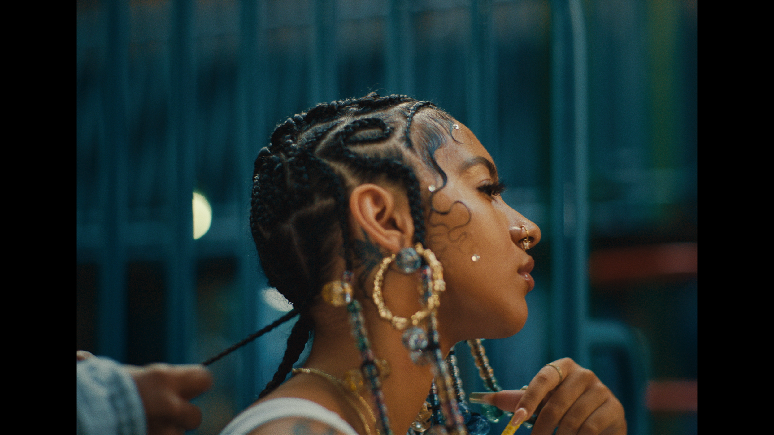 A woman with intricately braided hair adorned with beads, jewelry, and face piercings, viewed in profile with a blue background.