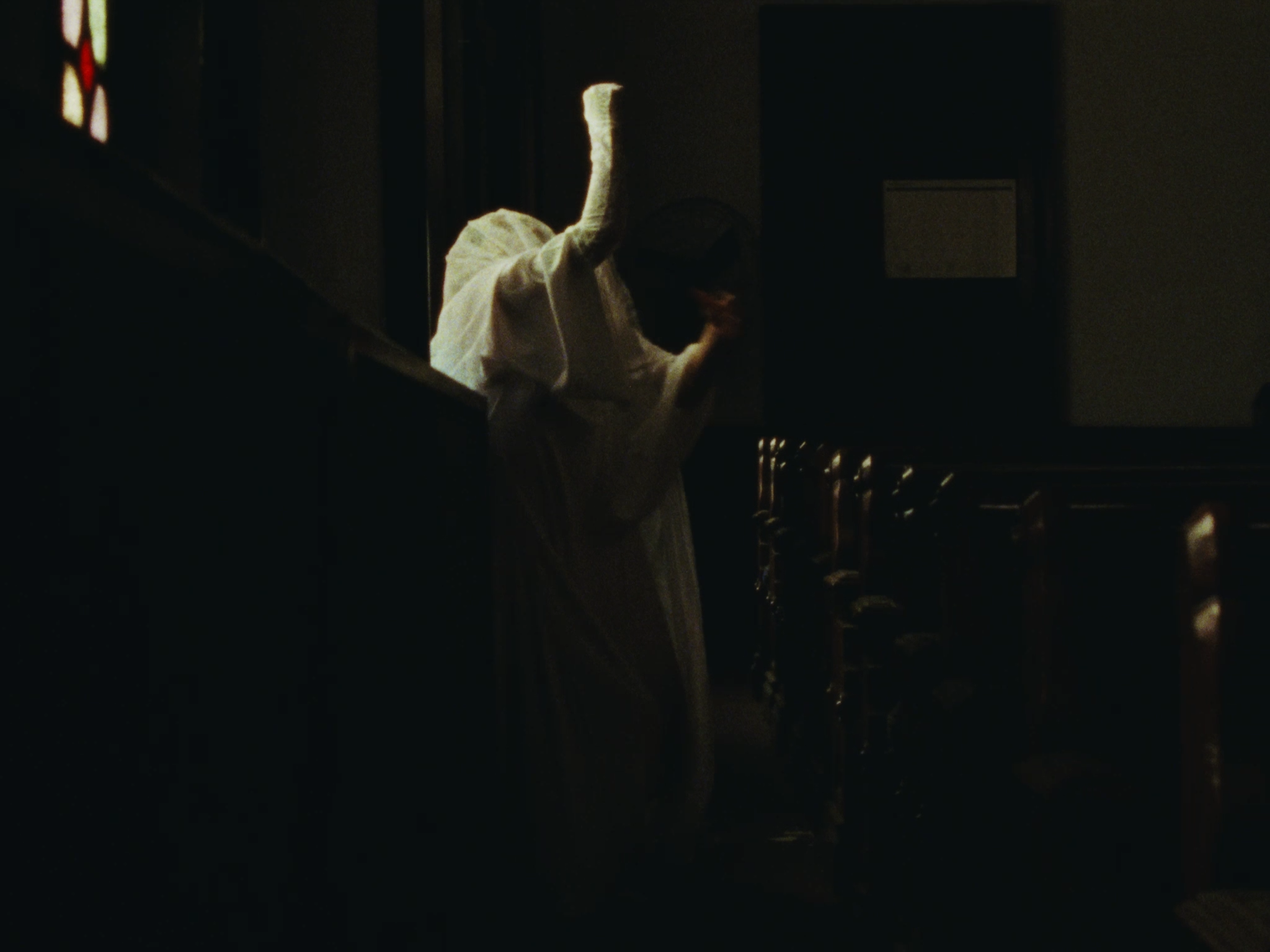 A person in a white robe and head covering bows in prayer inside a church, with stained glass windows and wooden pews visible in low lighting.