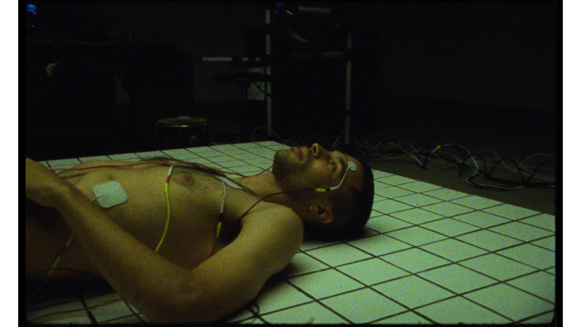 A man lying on a tiled floor with electrodes attached to his chest and a monitoring device on his wrist, in a dark room with medical or technical equipment in the background.