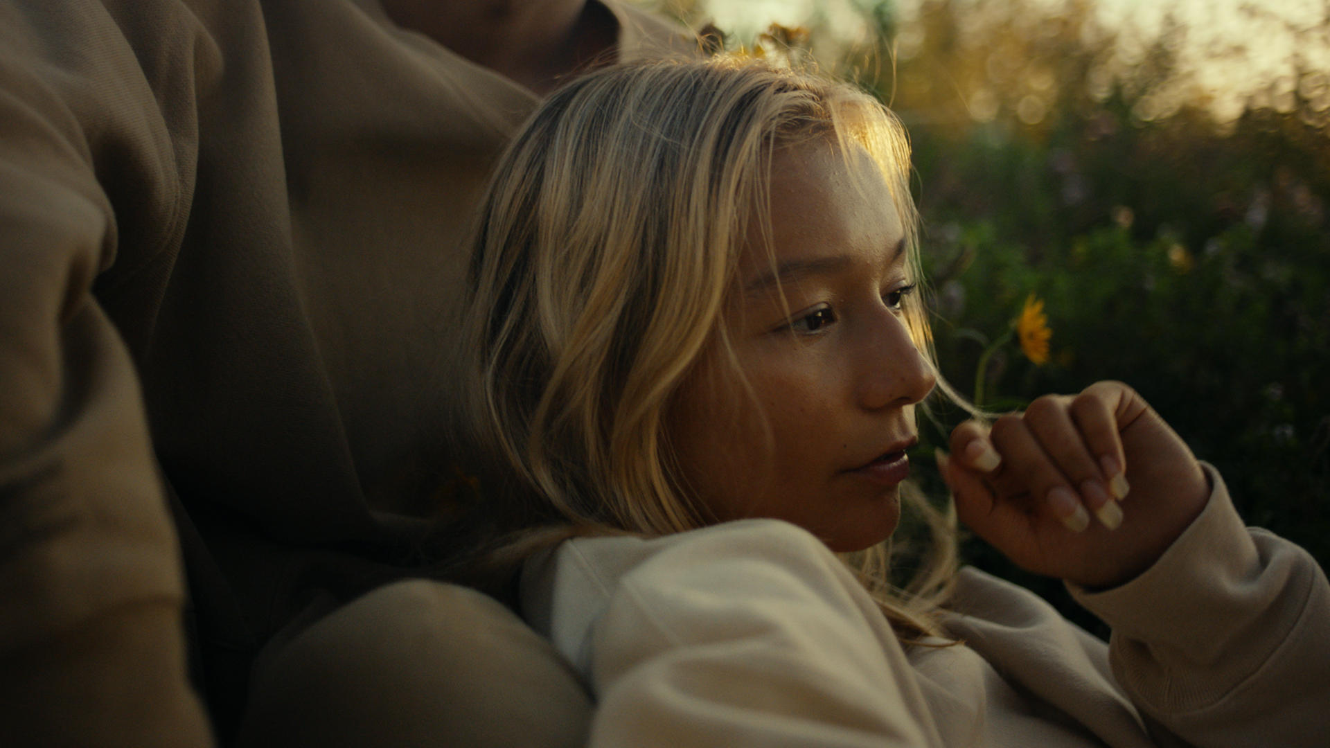 A young woman with blonde hair sitting outdoors near flowers, looking thoughtful with her hand near her face.