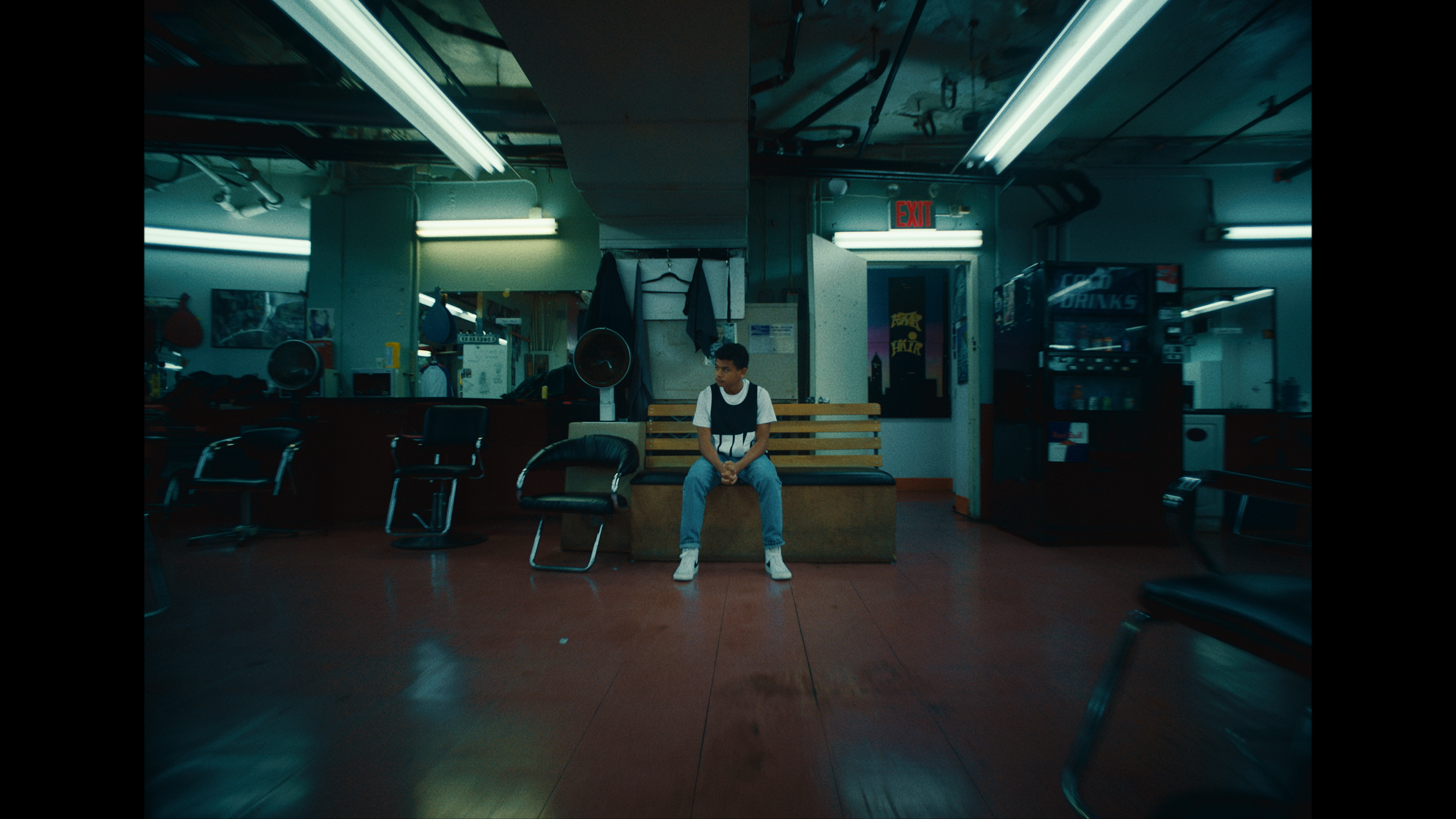 A young boy sitting alone on a bench in an empty, dimly lit cafe or restaurant, with chairs and vending machines around him.