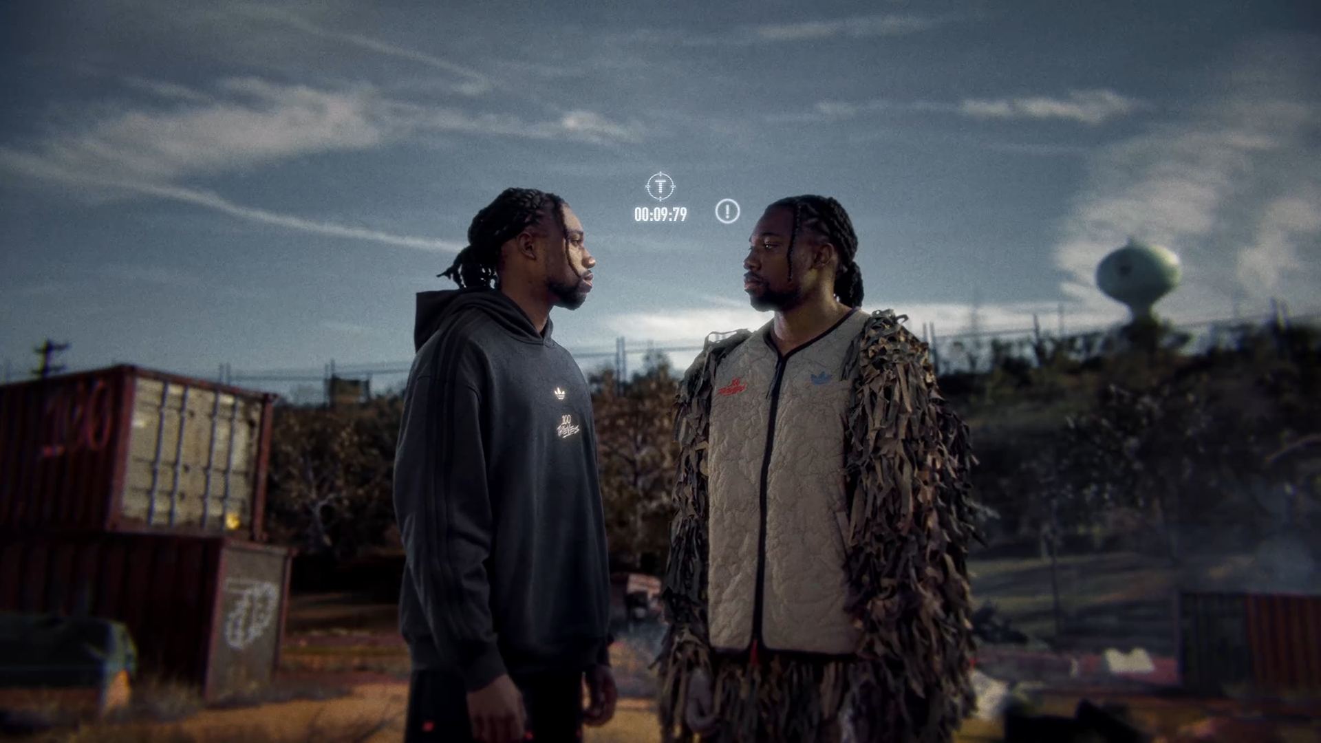 Two young men with braided hair standing face to face outdoors under a cloudy sky, with old shipping containers and a water tower in the background.
