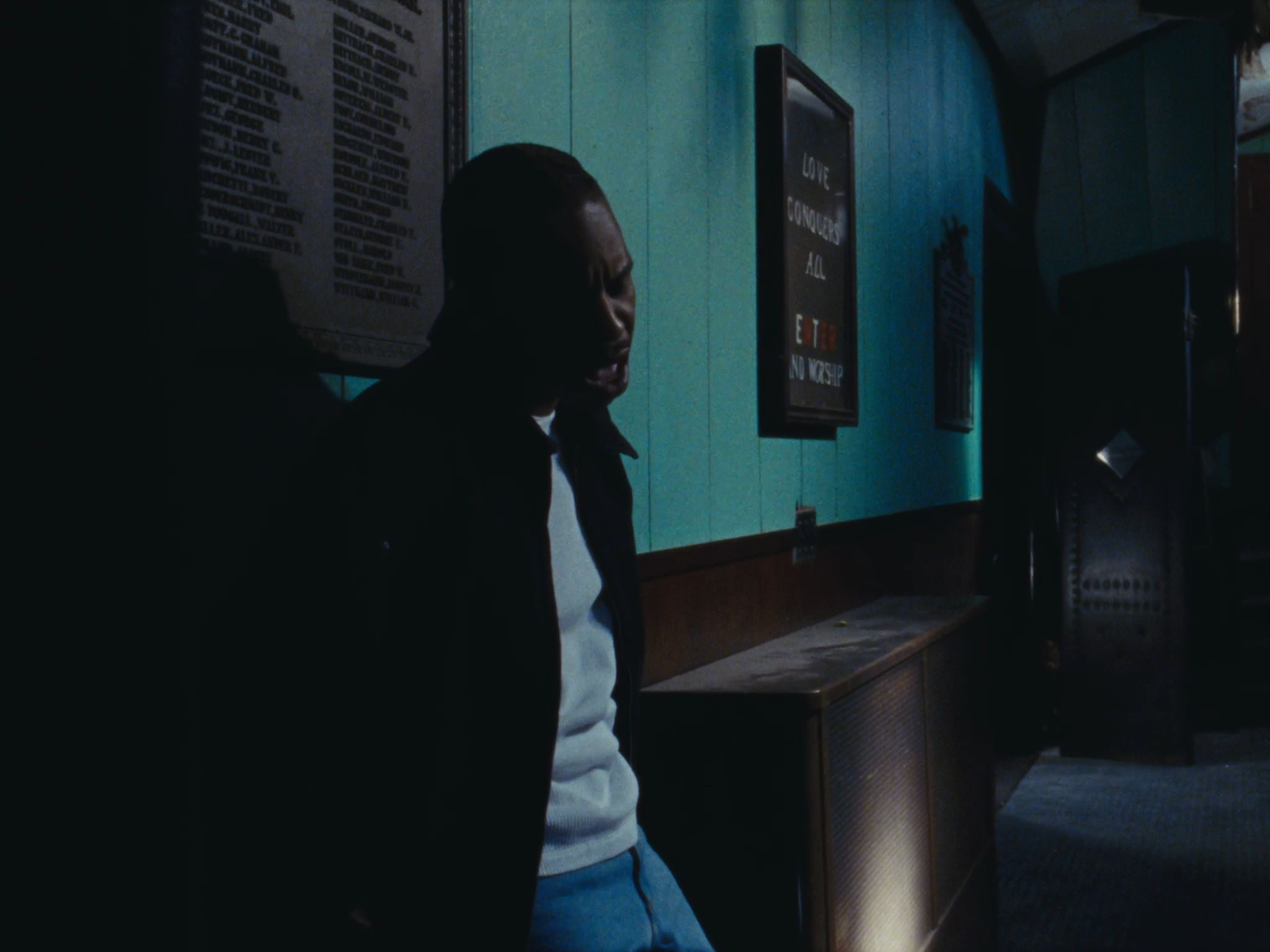 A young man standing in a dimly lit room with blue walls, looking down, with framed posters on the wall, one of which reads 'Love Conquered All, E To No Worship'.