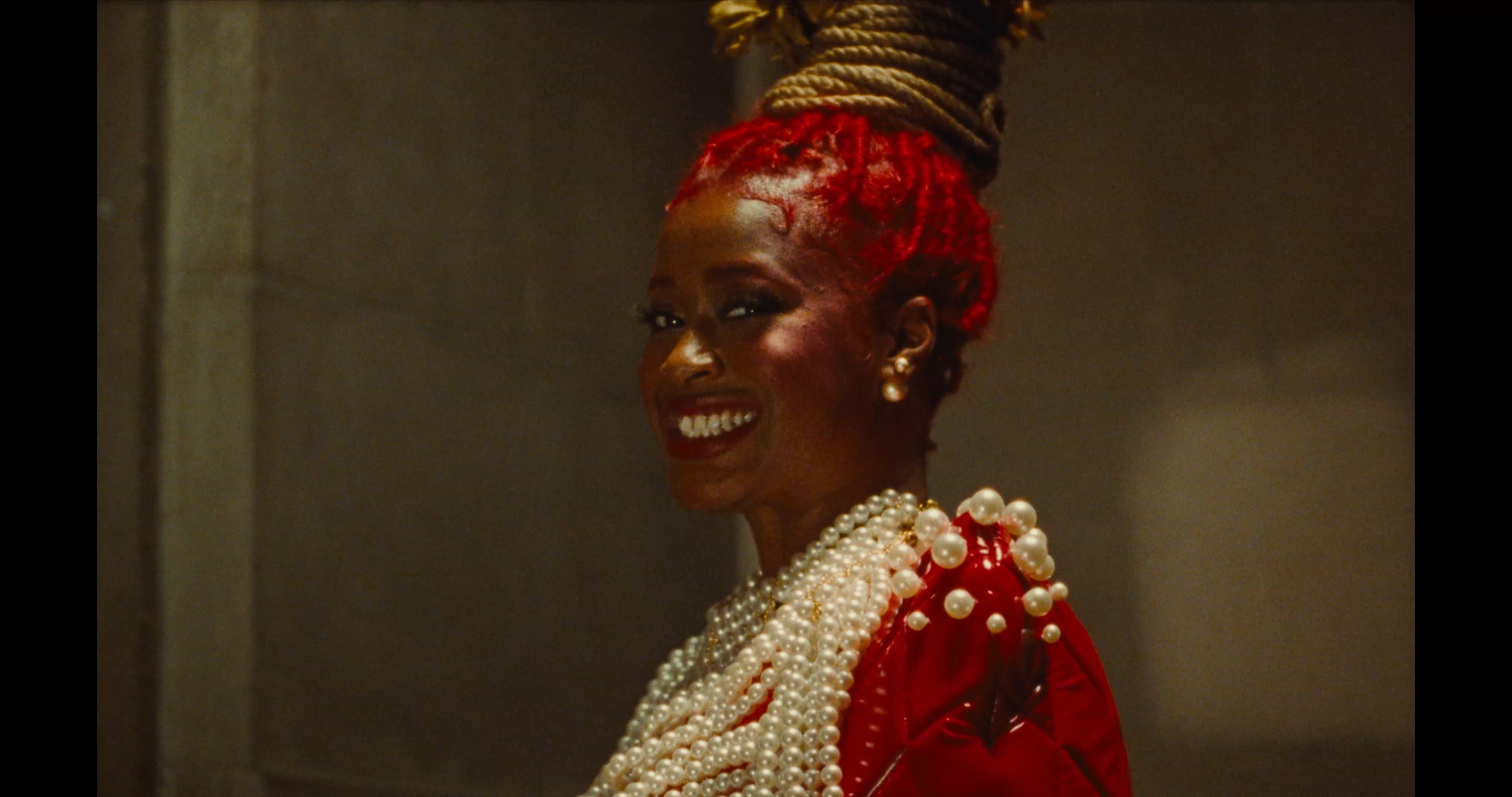 A smiling woman with red braided hair styled in an updo, wearing pearl jewelry and a red outfit, standing against a plain background.