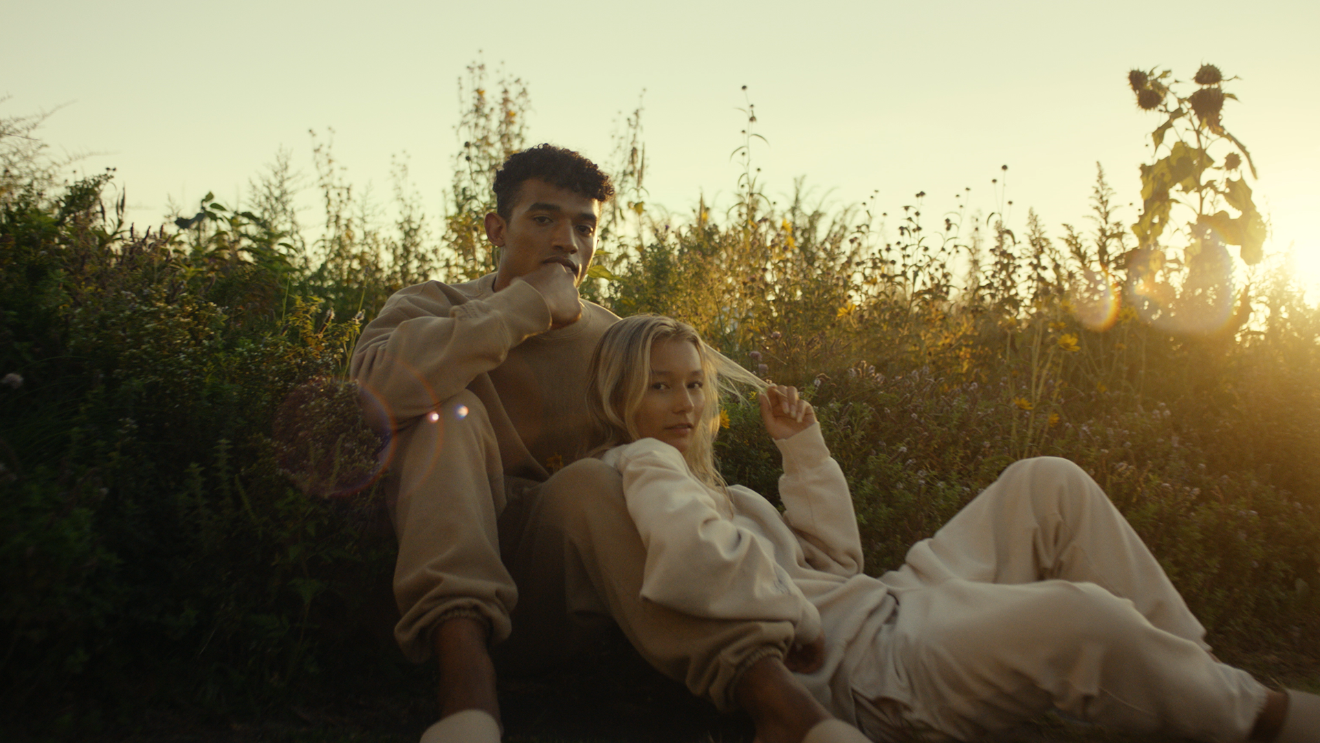 A young man and woman sitting on the ground in a field of tall plants during sunset. The man has curly hair and is wearing a beige sweatshirt, and the woman has blonde hair and is wearing a light-colored outfit. The woman is reclining with her head o