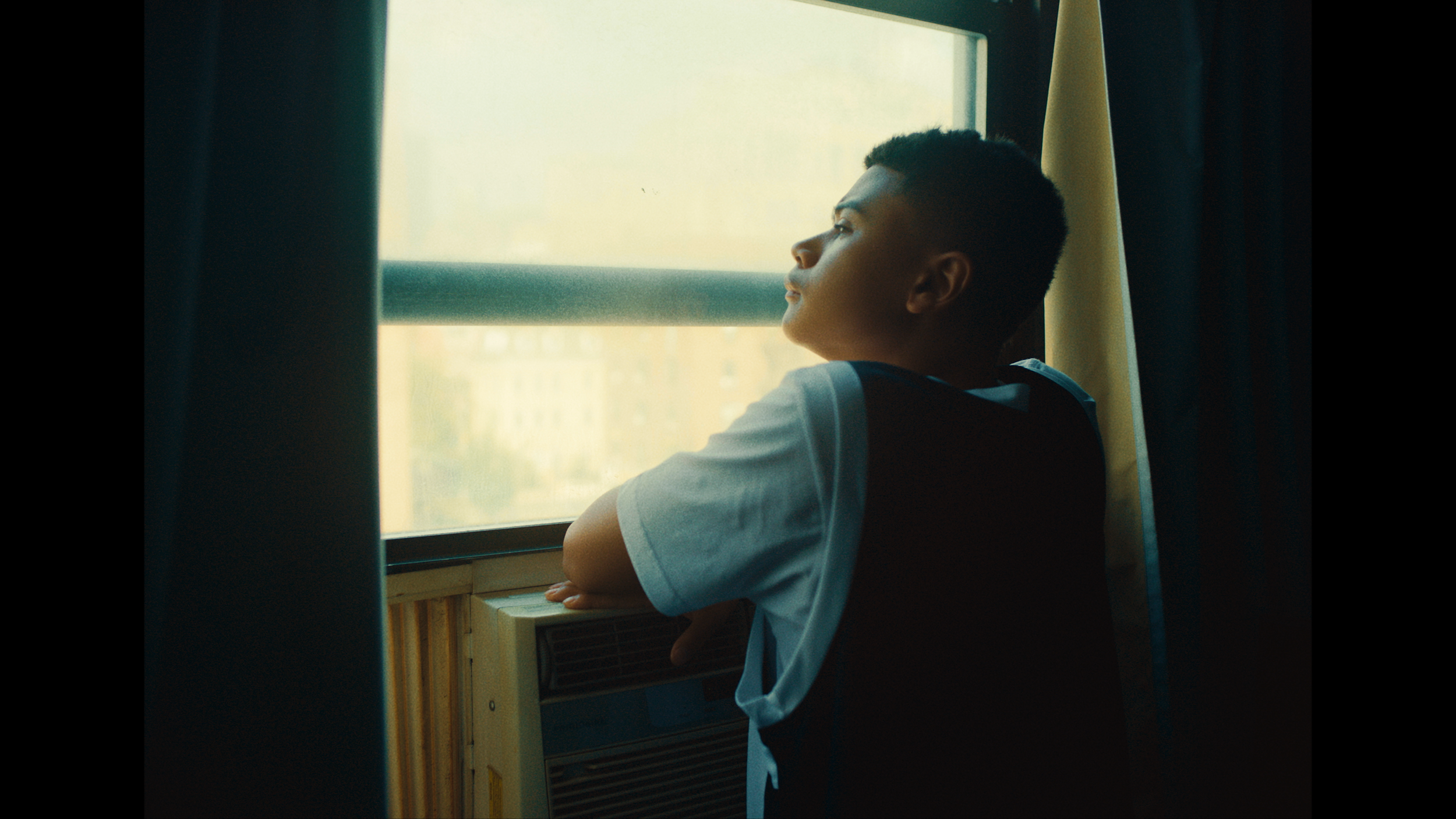 A young boy wearing a white T-shirt and dark vest looking out a bus window, resting his arms on the window ledge, with sunlight illuminating his face.