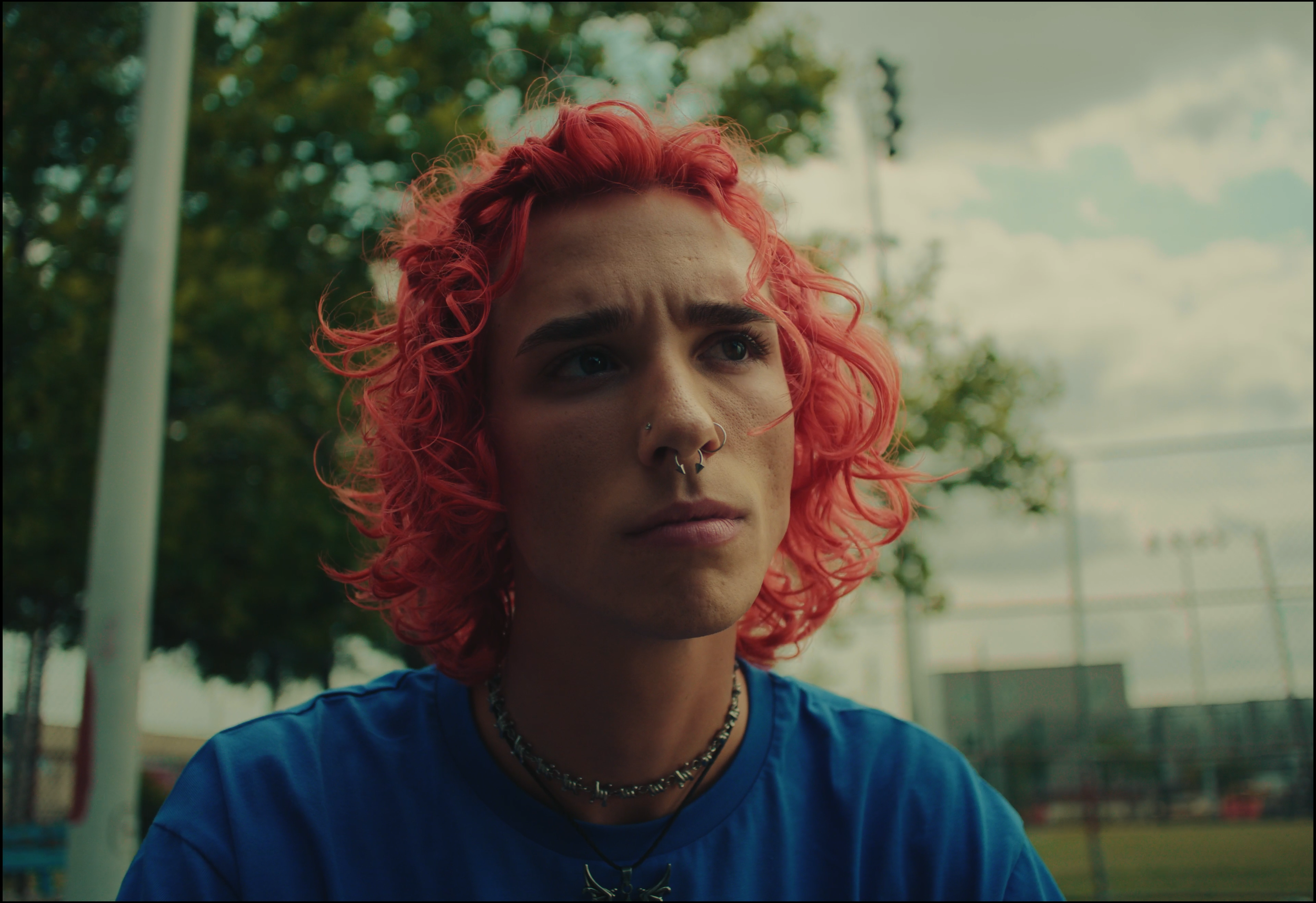 A young woman with curly pink hair, wearing a dark choker and a blue shirt, gazes thoughtfully outdoors with trees and a sports field in the background.