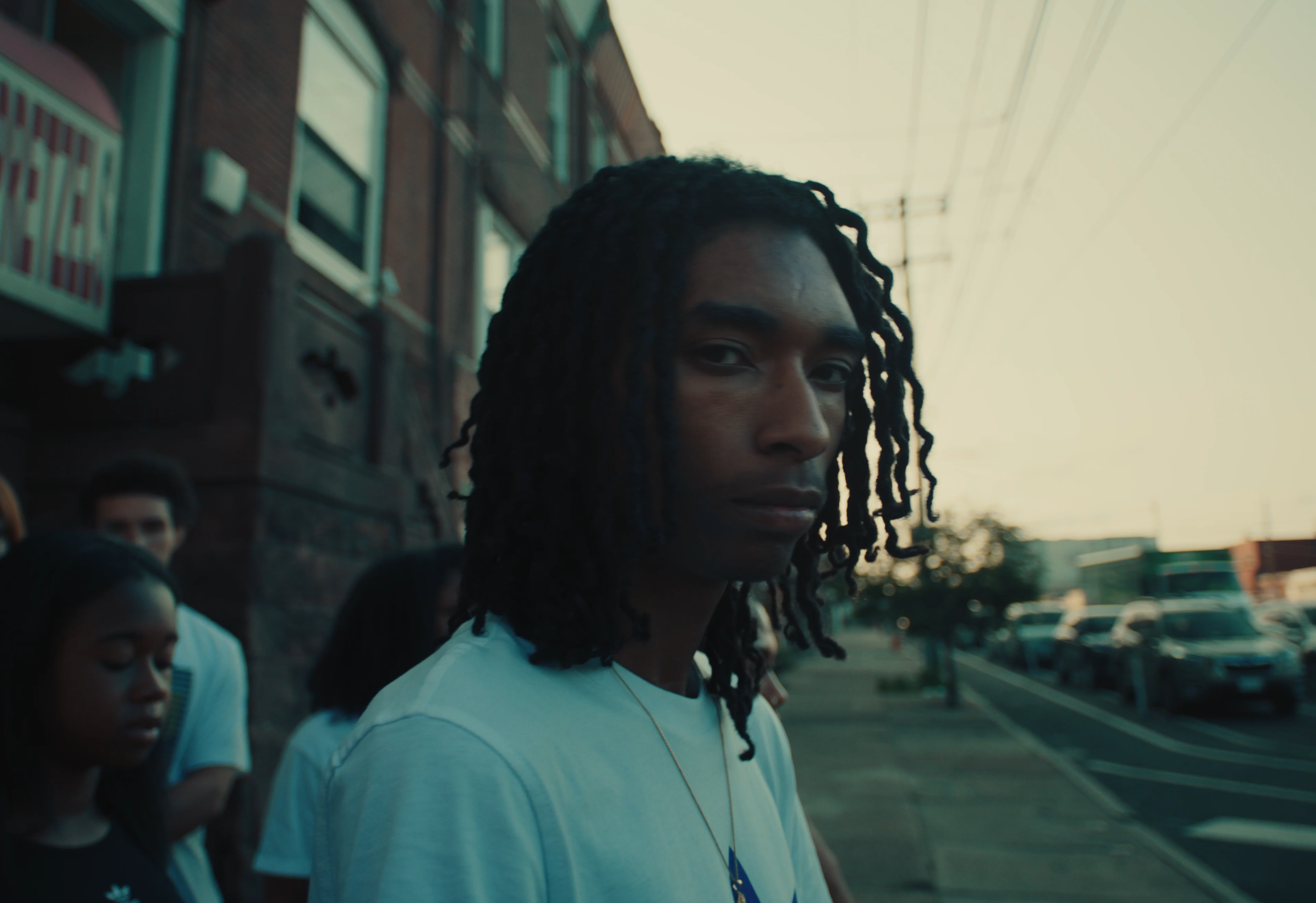 A young woman with shoulder-length dreadlocks and a serious expression standing outside during sunset, with a brick building and parked cars in the background, alongside other people.