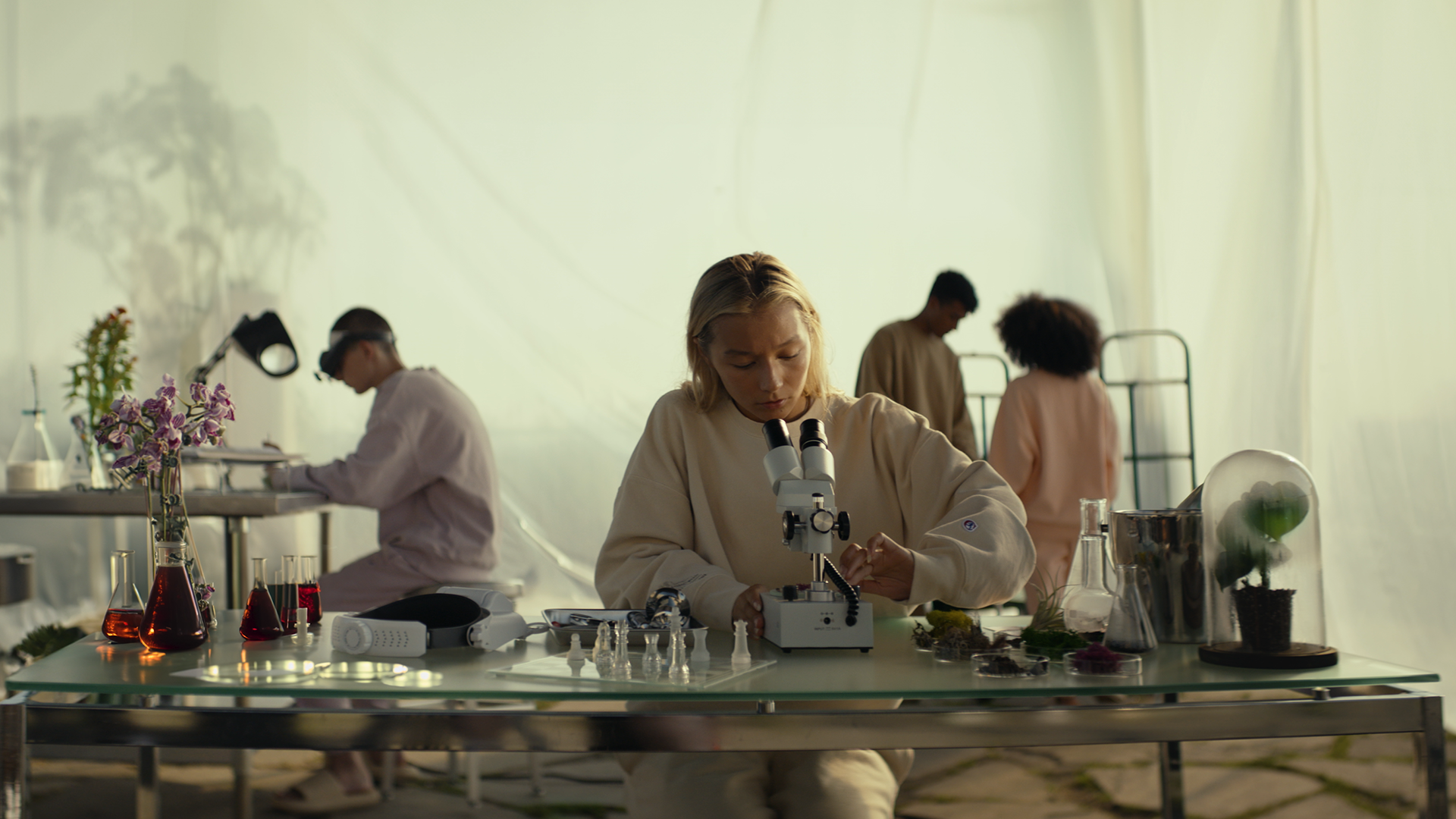 A woman looking through a microscope at a laboratory table with plants and glassware, with three people working in the background.