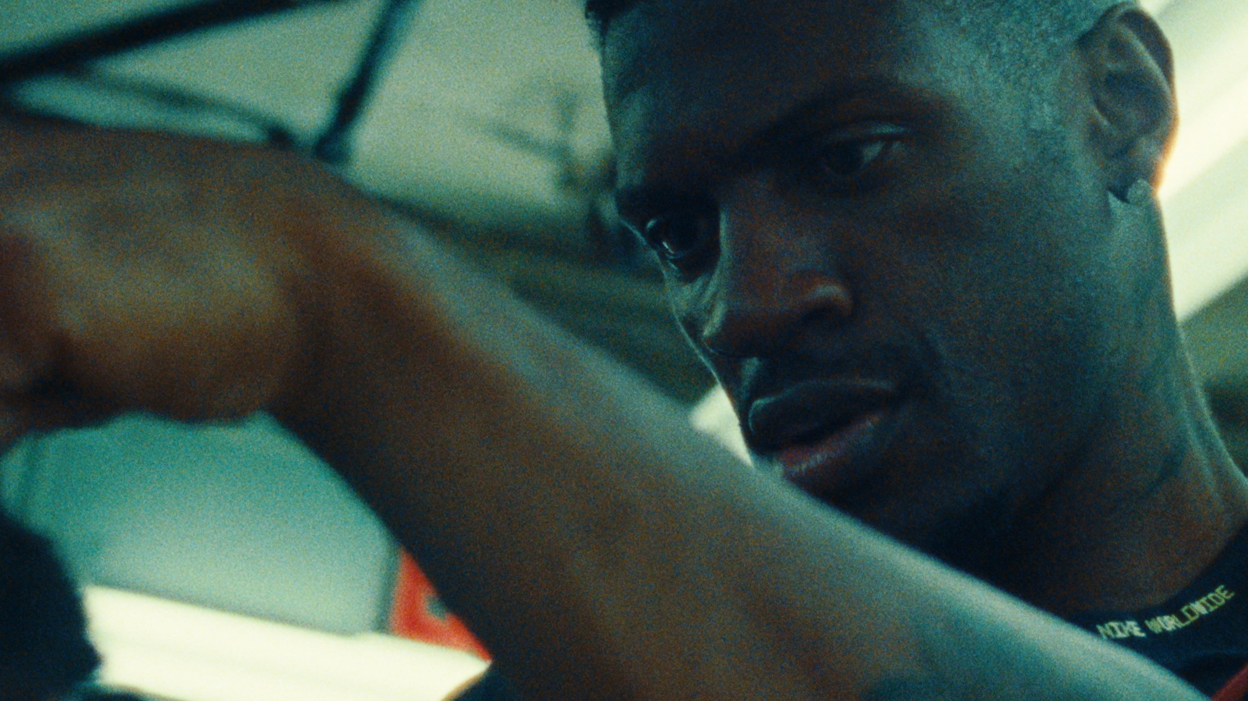 A young man with dark skin and short hair is looking over his shoulder, with a serious expression, in a close-up shot. The background appears to be indoors with a muted color palette.