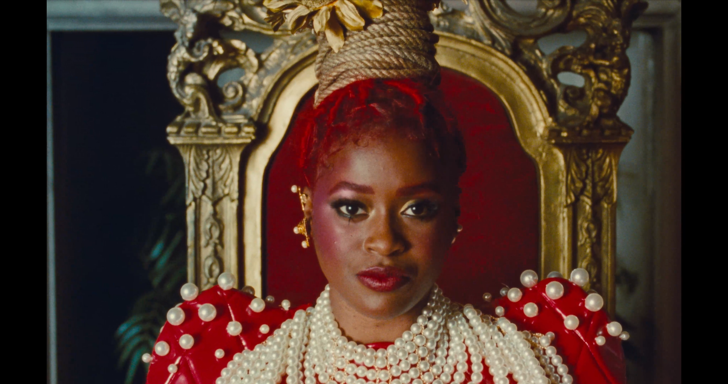 A woman with red hair styled in an updo, sitting on a grand ornate golden throne with a red velvet cushion. She is wearing a red dress with pearl embellishments and multiple strands of pearls around her neck, along with earrings. The background featu