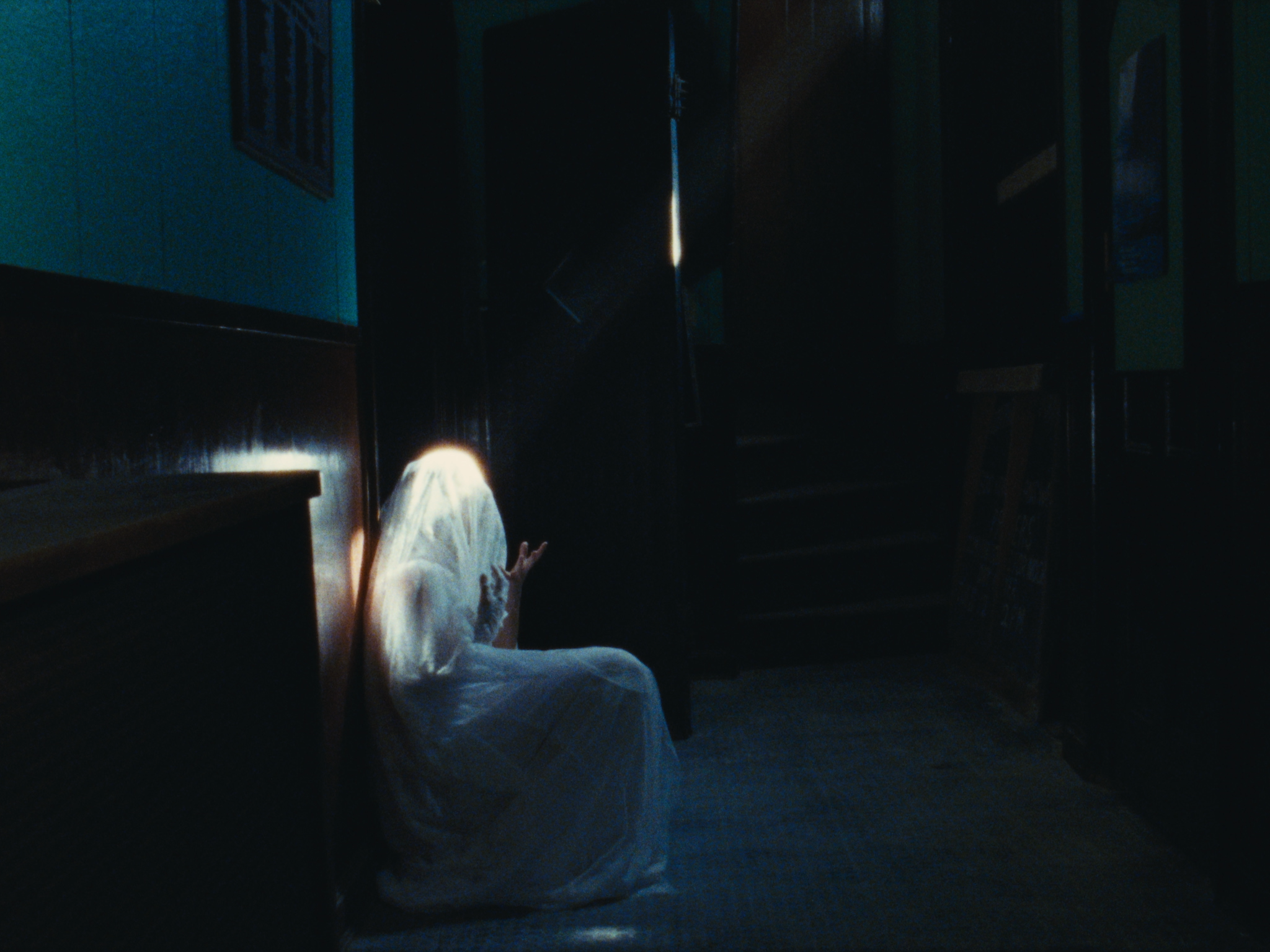 A ghostly figure with a white sheet and hand raised is sitting on the floor in a dark hallway.