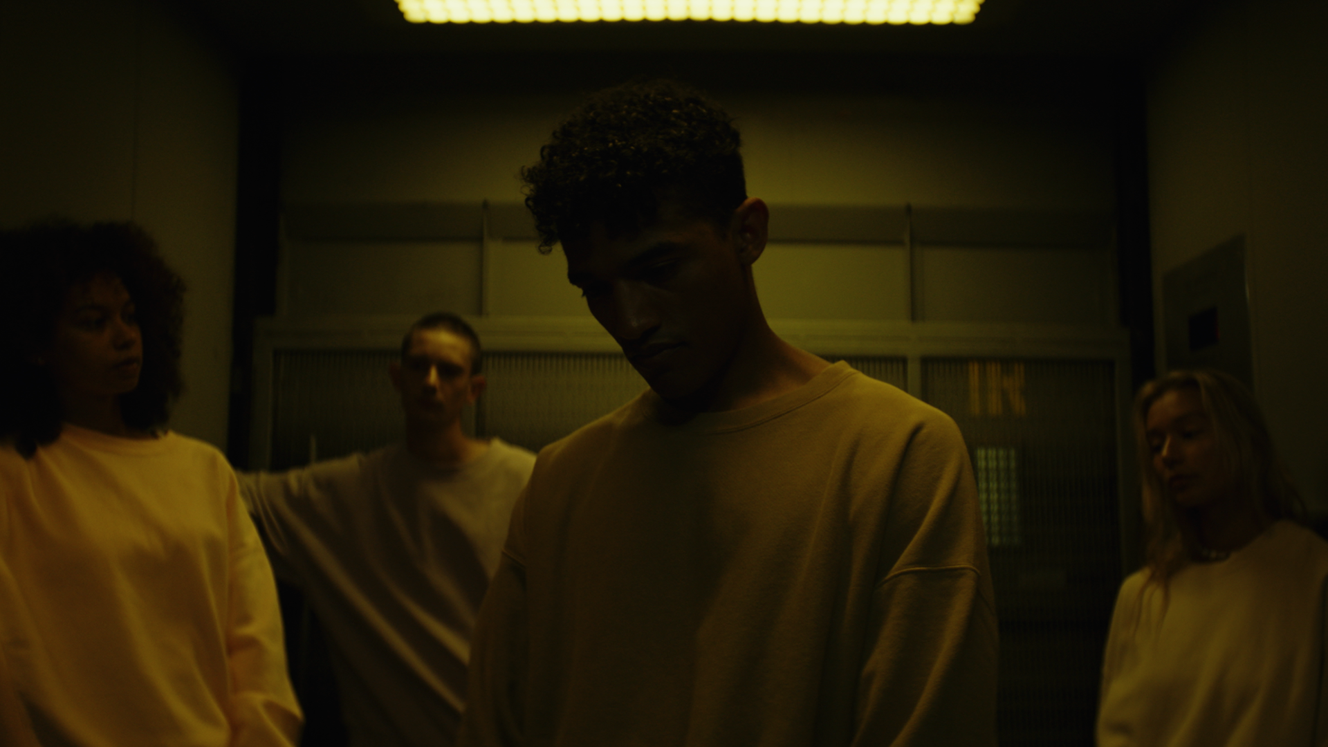 Four young people standing in a dimly lit room, with one person in the foreground and three behind, all wearing light-colored clothing.