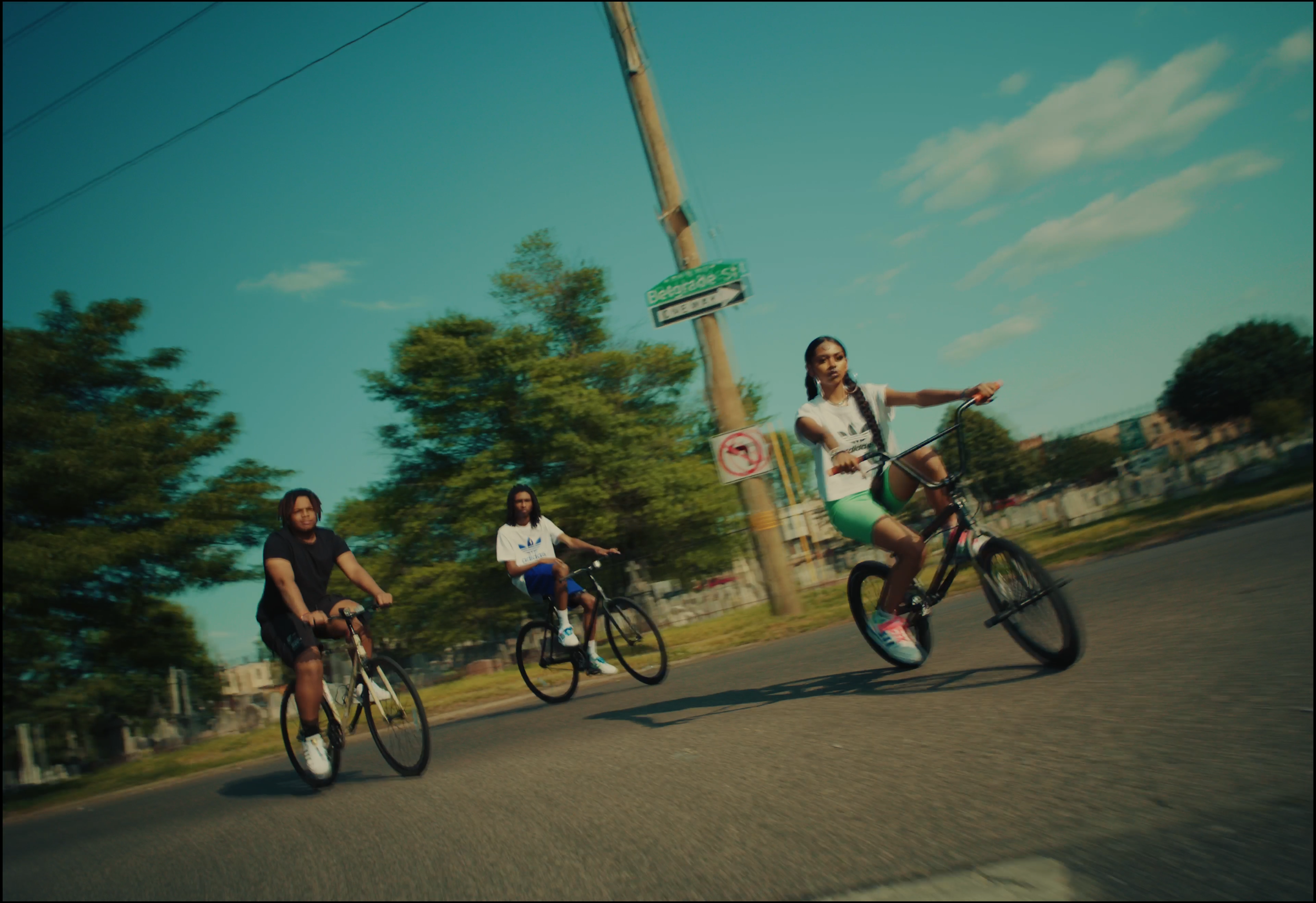 Three young women riding bicycles on a city street with trees and a utility pole with street signs in the background