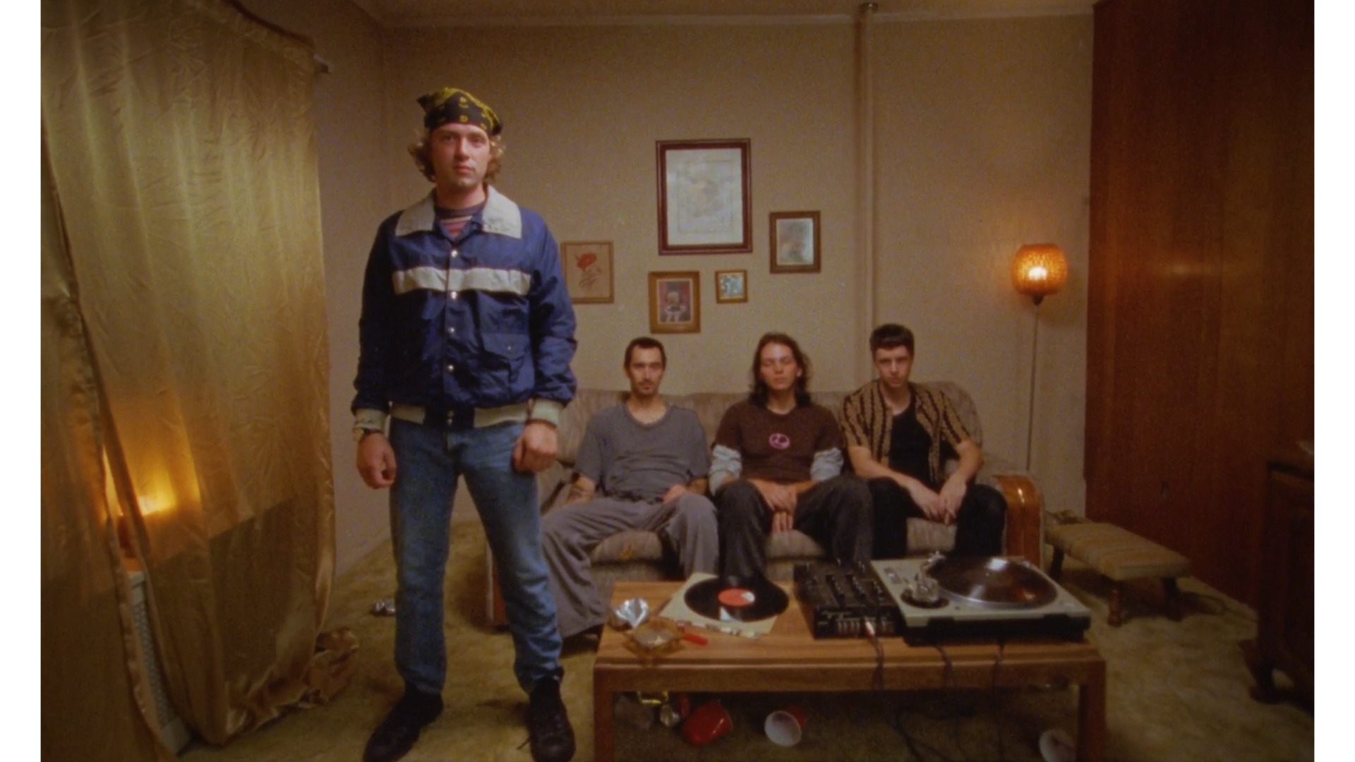 Four young adults in a living room, three are sitting on a couch and one is standing in front of them. The room has vintage decor, a coffee table with records and tech equipment, and a lit wall lamp.