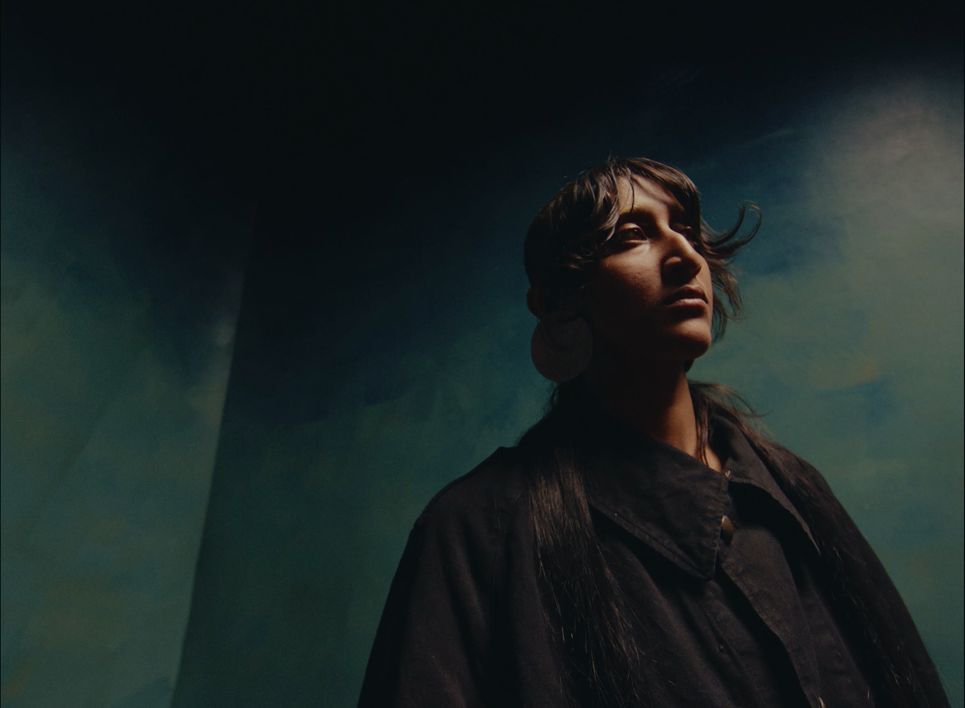 A woman with long dark hair in a dark jacket gazes upward with a contemplative expression, against a dark, abstract background with hints of green and blue.