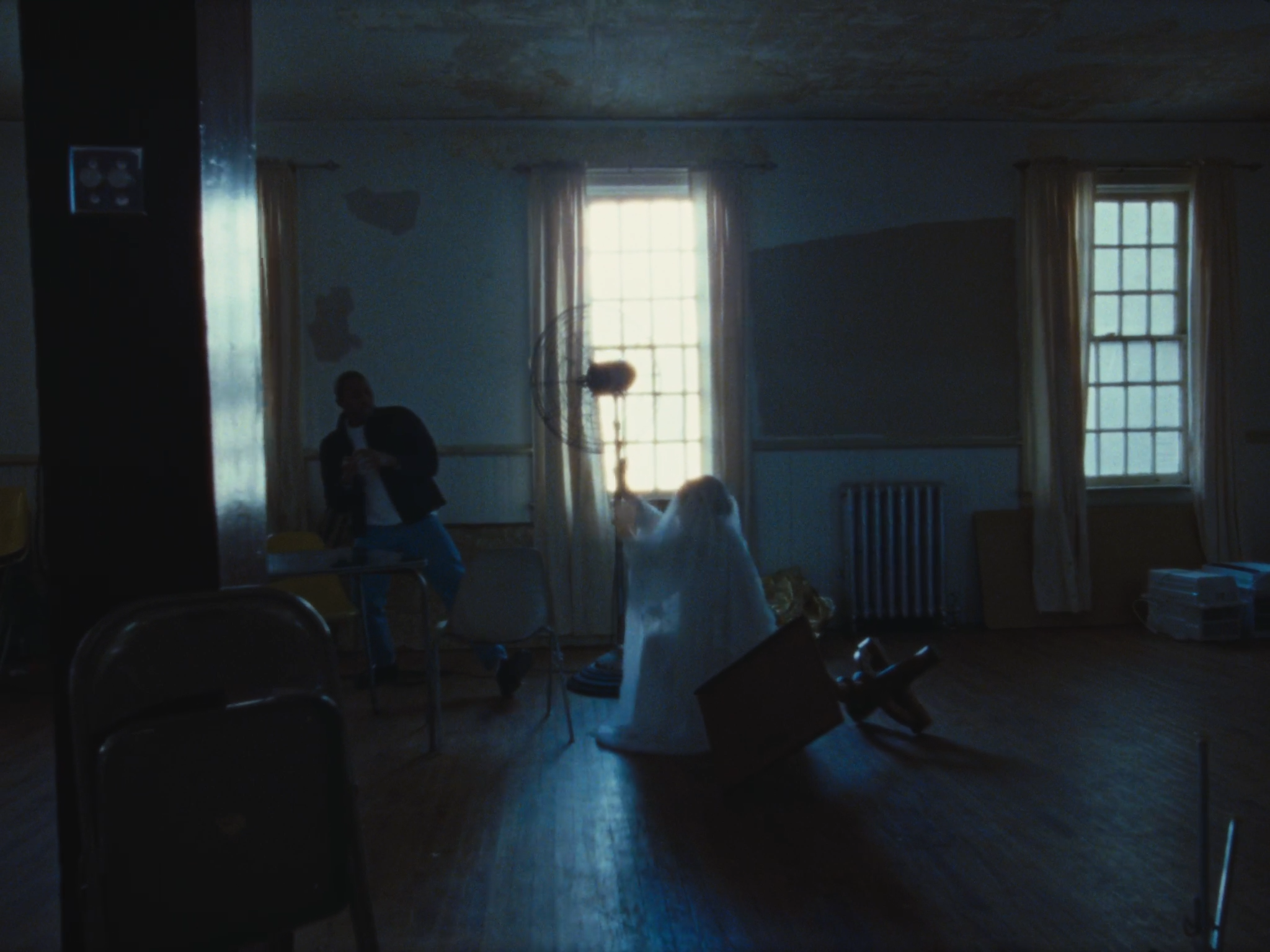 Dimly lit room with two windows and curtains, a person with long hair sitting on the floor, a person standing near the window, a table, and some chairs.